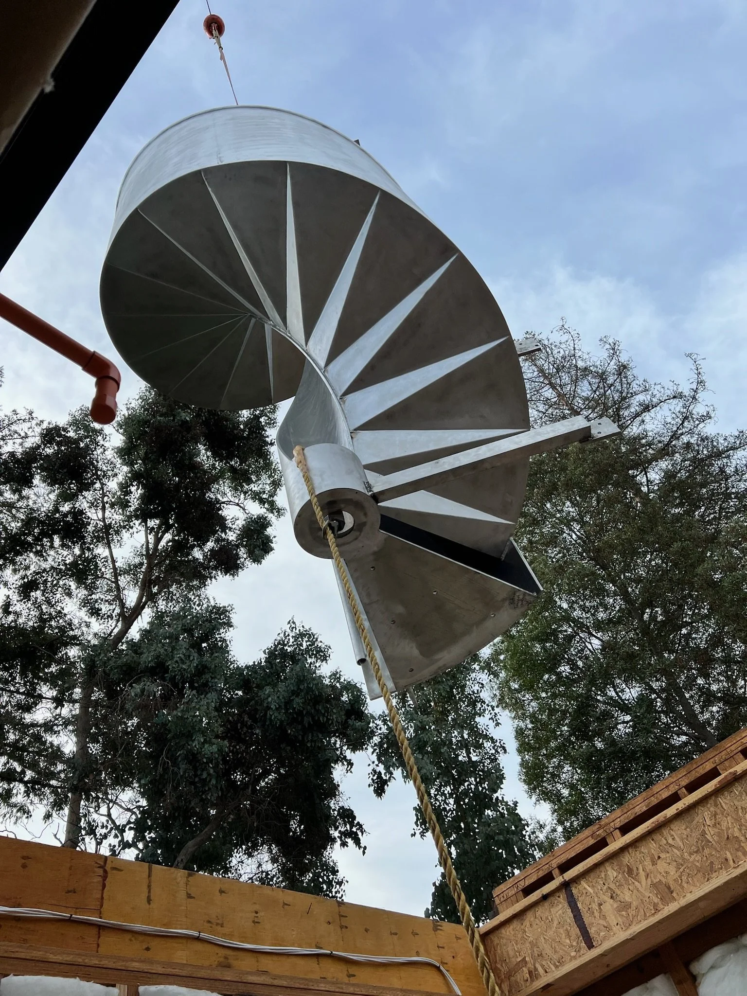 A spiral staircase made of metal with wide steps and a cylindrical central support, viewed from below against a cloudy sky with trees in the background.