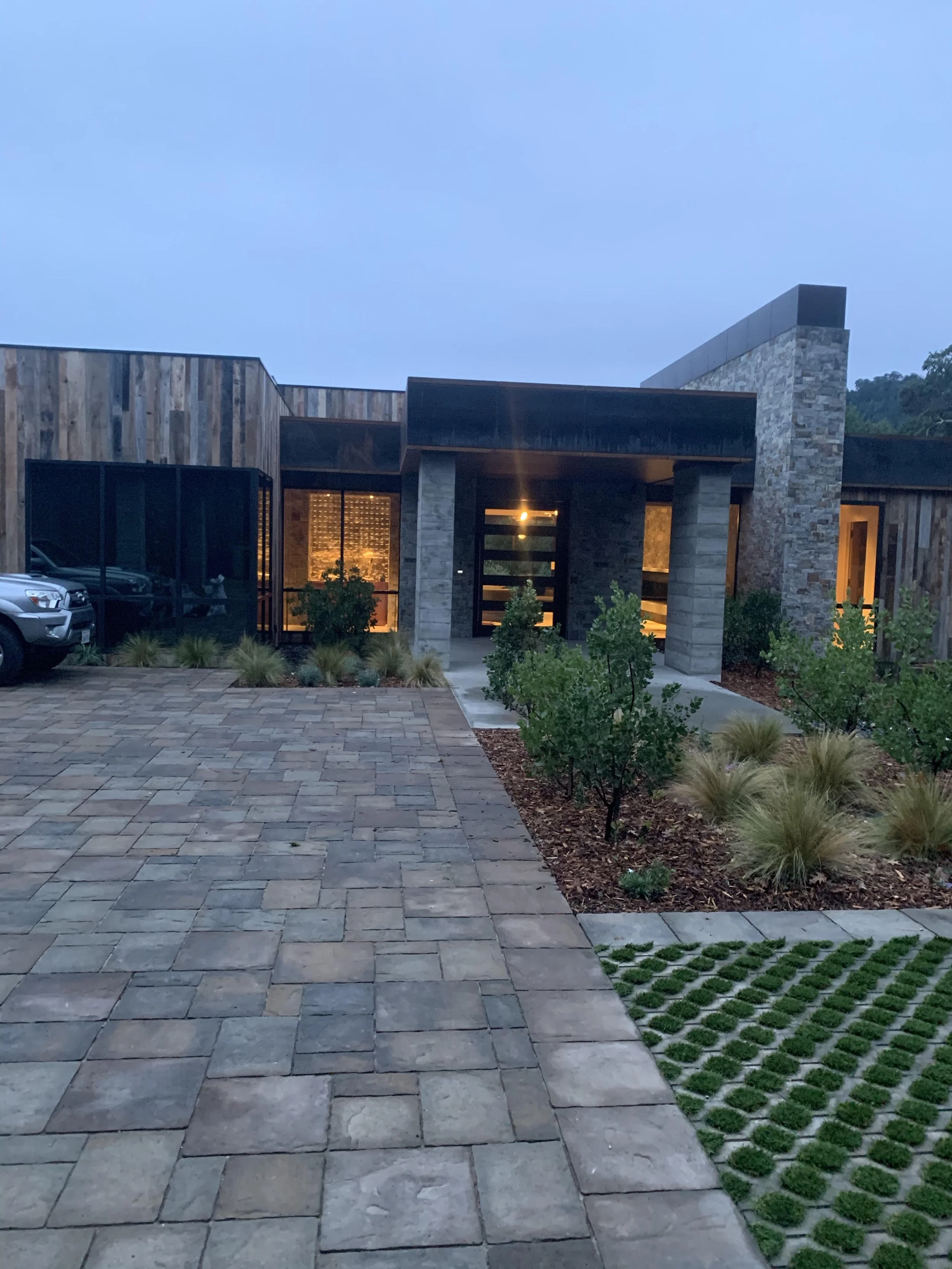 Modern house with a stone and wood exterior, lit interior lights visible through windows, a paved driveway, and landscaped bushes and grass in front.
