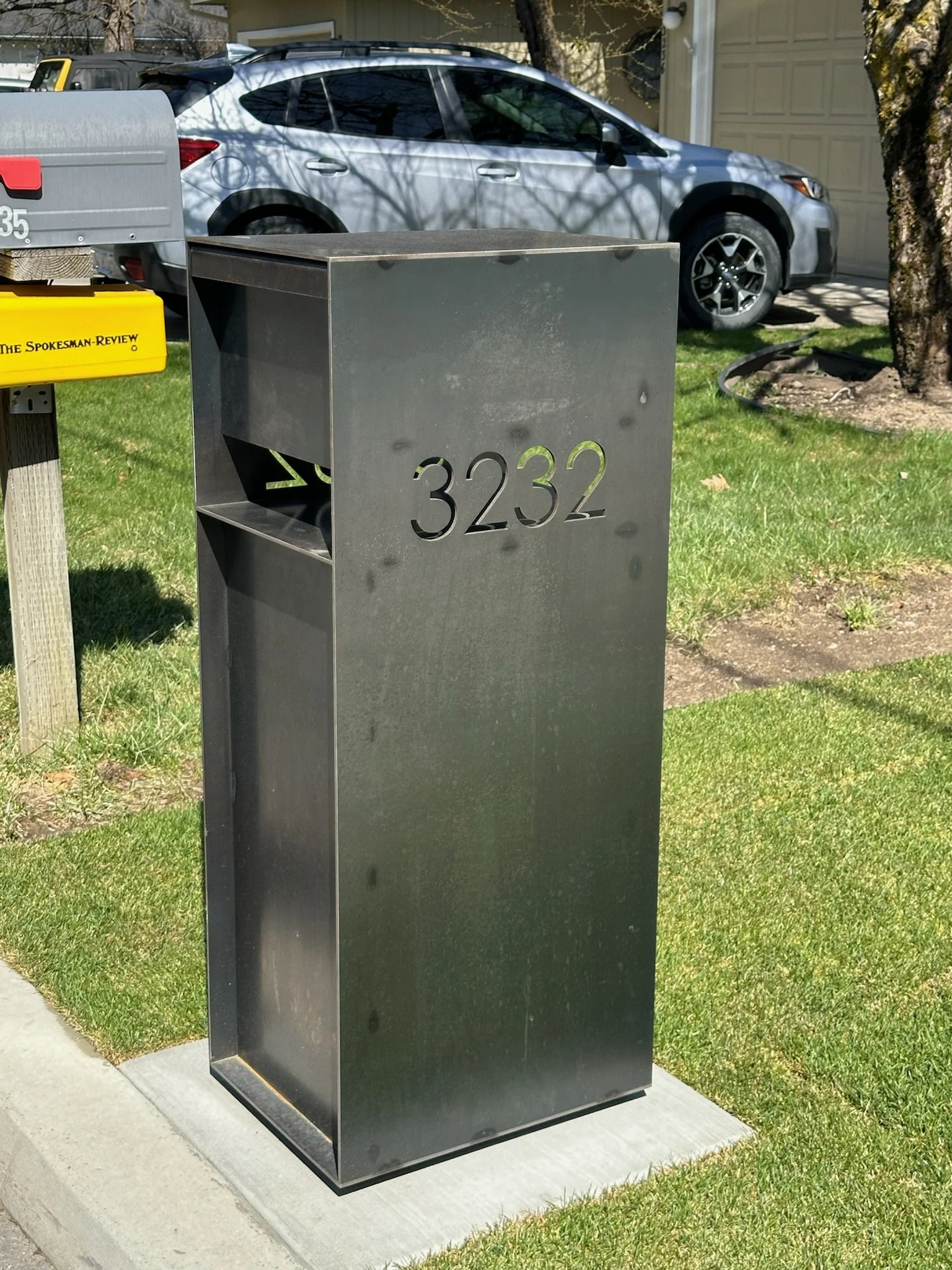 Black metal mailbox with the house number 3232. It is placed on a concrete slab next to a sidewalk, with a green lawn and a tree nearby. A silver car is parked in the driveway in the background, along with a yellow mail collection box.