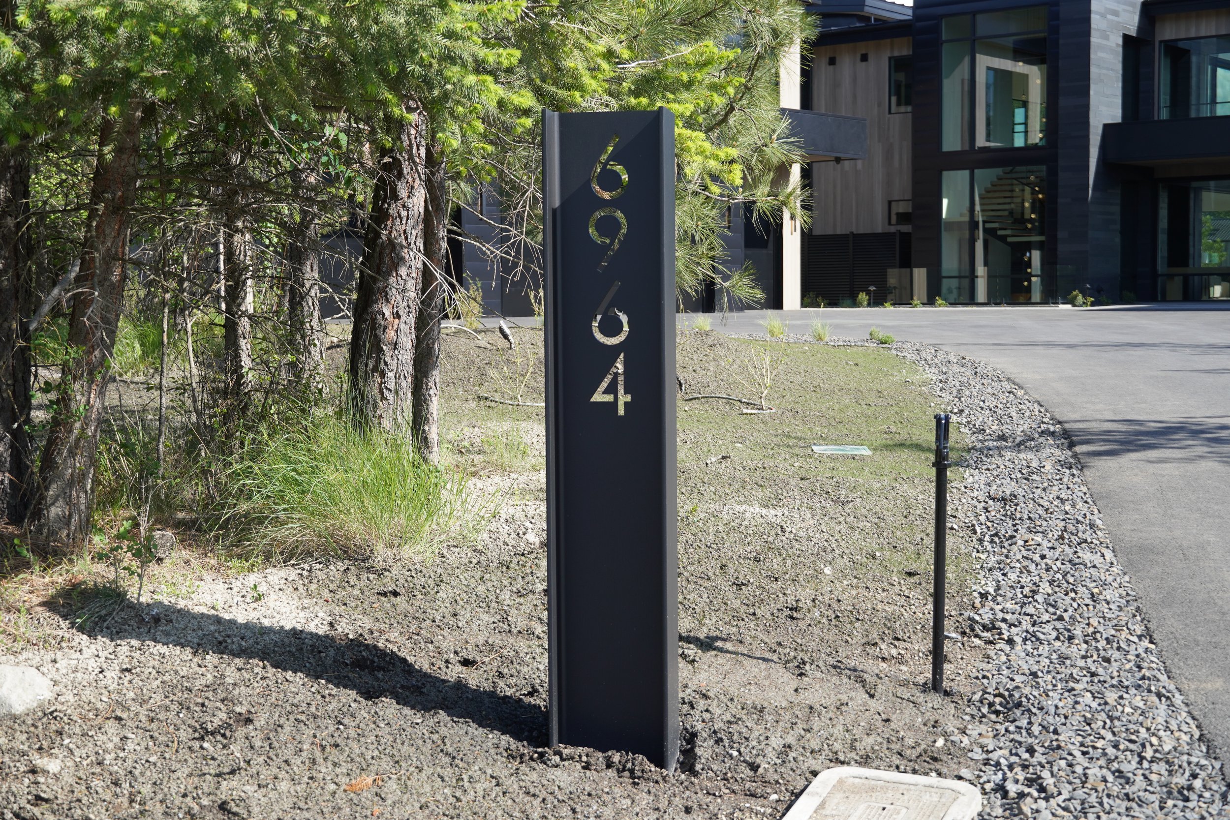 Modern building withlarge windows and stairs, trees and shrubs nearby, metal signpost with numbers, gravel and concrete pavement, lush greenery in the background.