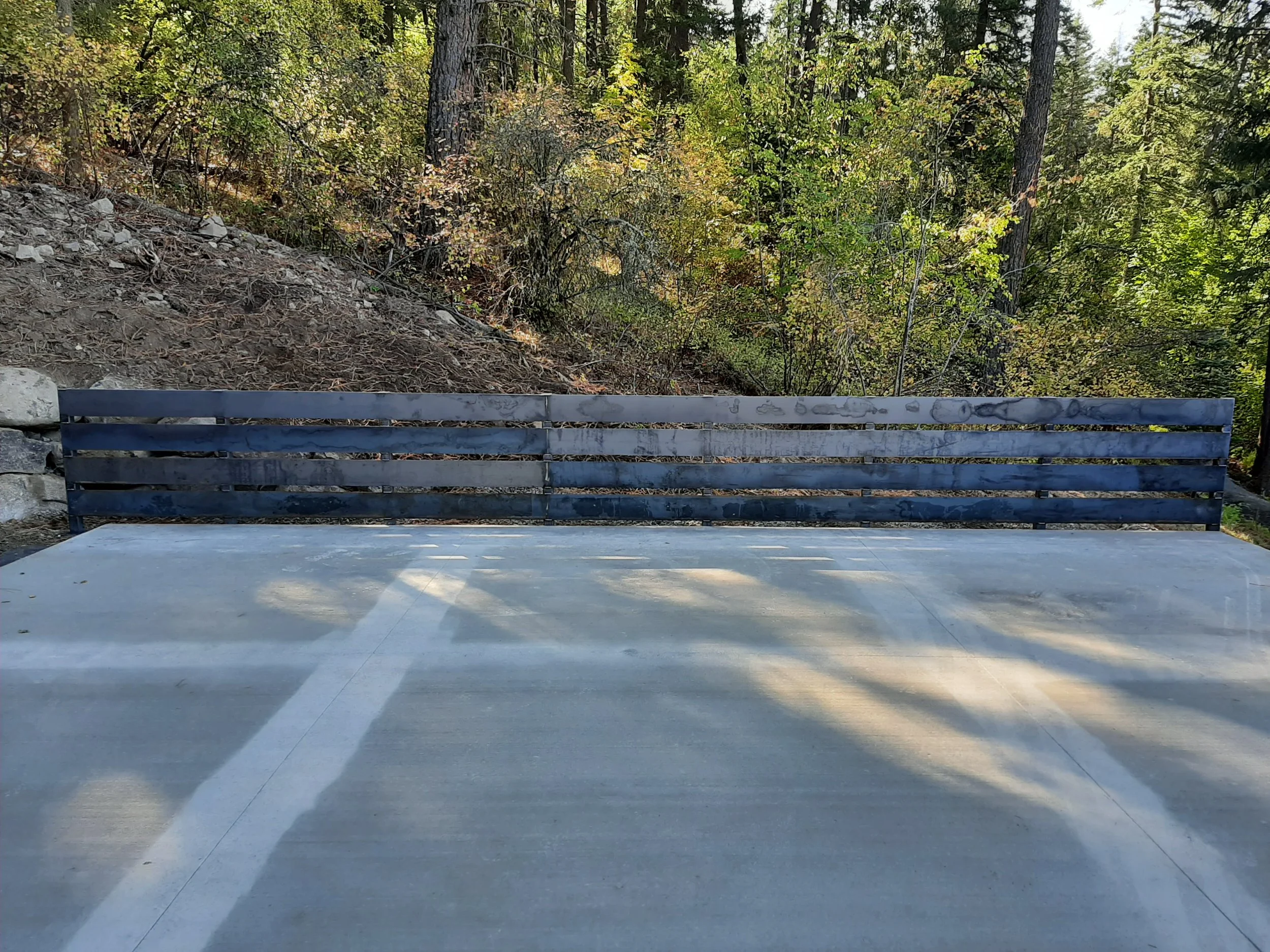 A bench with a wooden backrest and a concrete seat located outdoors on a concrete surface, with a wooded area in the background.