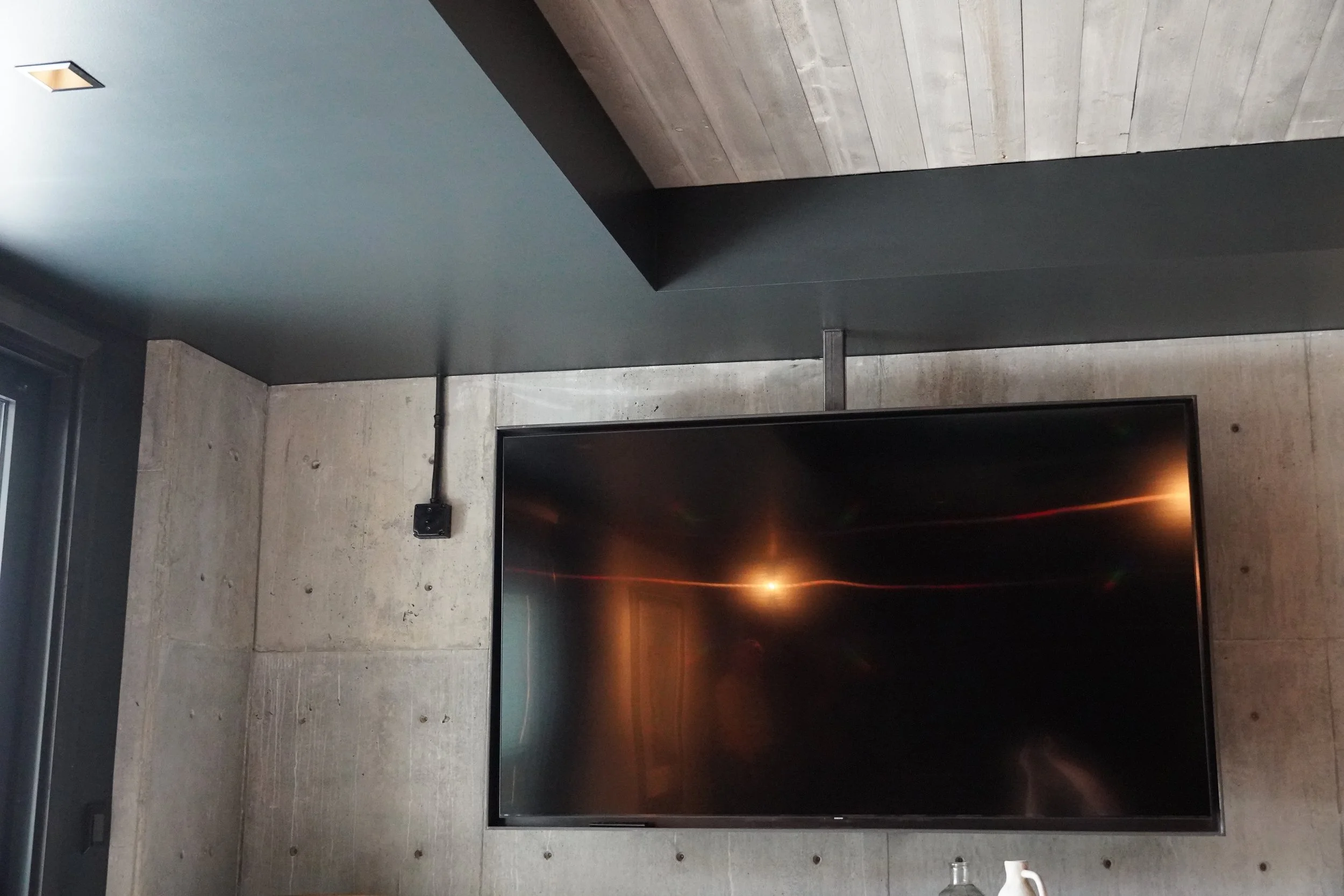 Interior of a modern room with concrete walls, a large flat-screen TV, a ceiling with wood paneling, and a window on the left side.