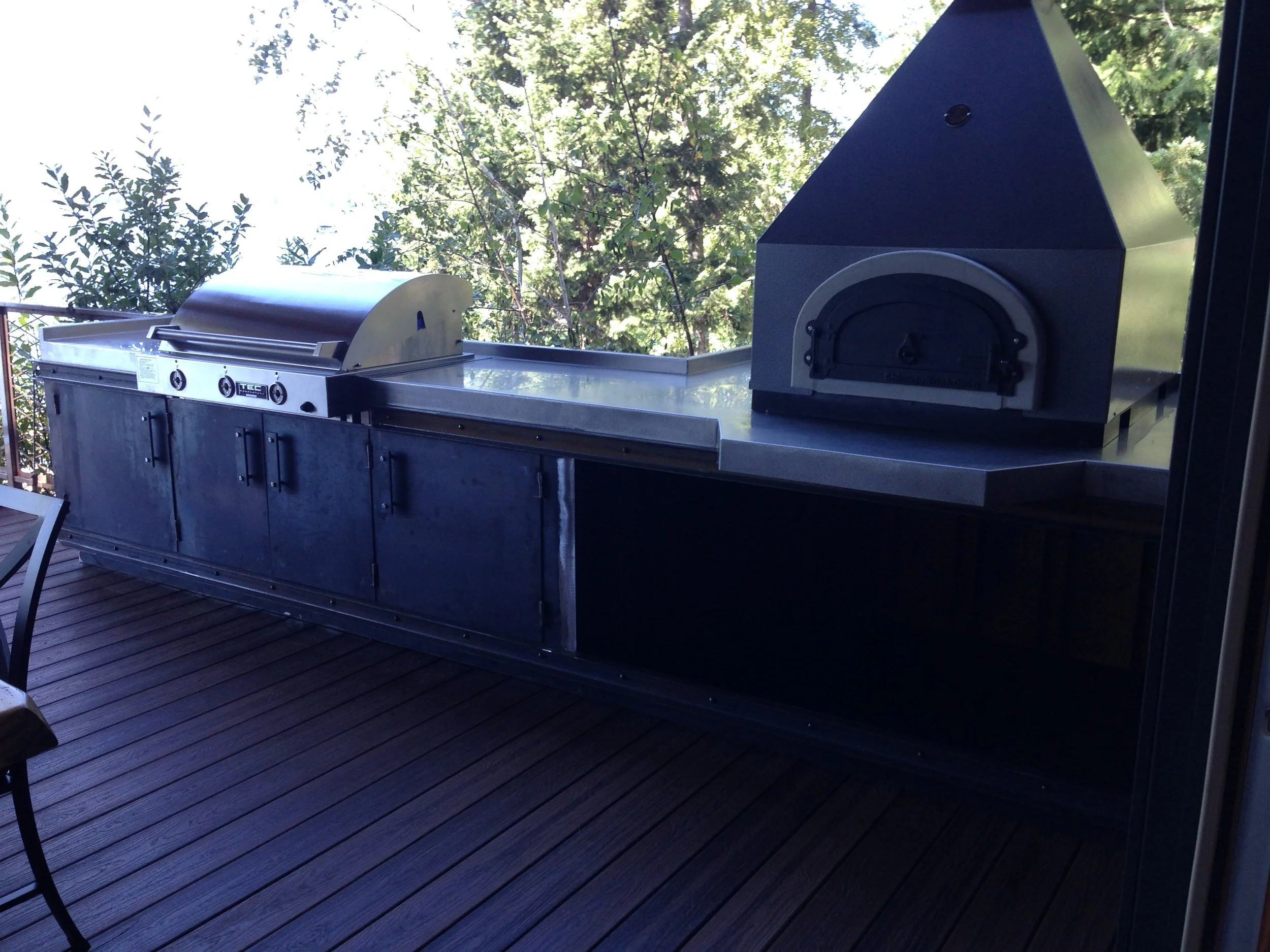 Outdoor patio with two stainless steel grilling appliances and a wooden deck, surrounded by trees and greenery, with partly cloudy sky in the background.