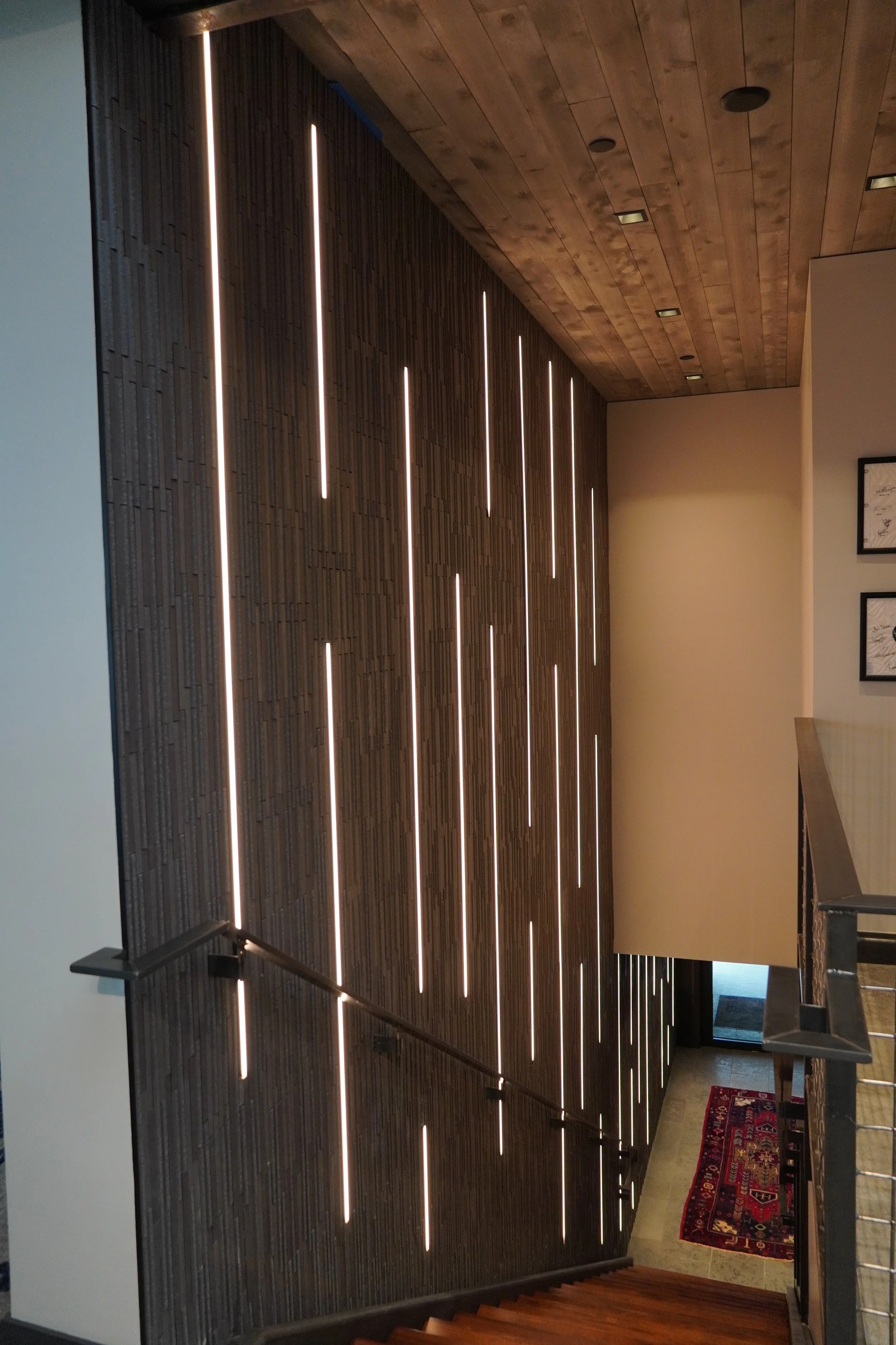 Modern staircase with a wooden wall featuring vertical LED light strips, leading down to a small landing with a door and a red patterned rug.