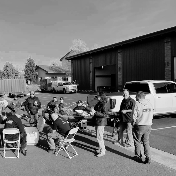 People gathered outside a fire station, serving food and drinks, with trucks and a building in the background.
