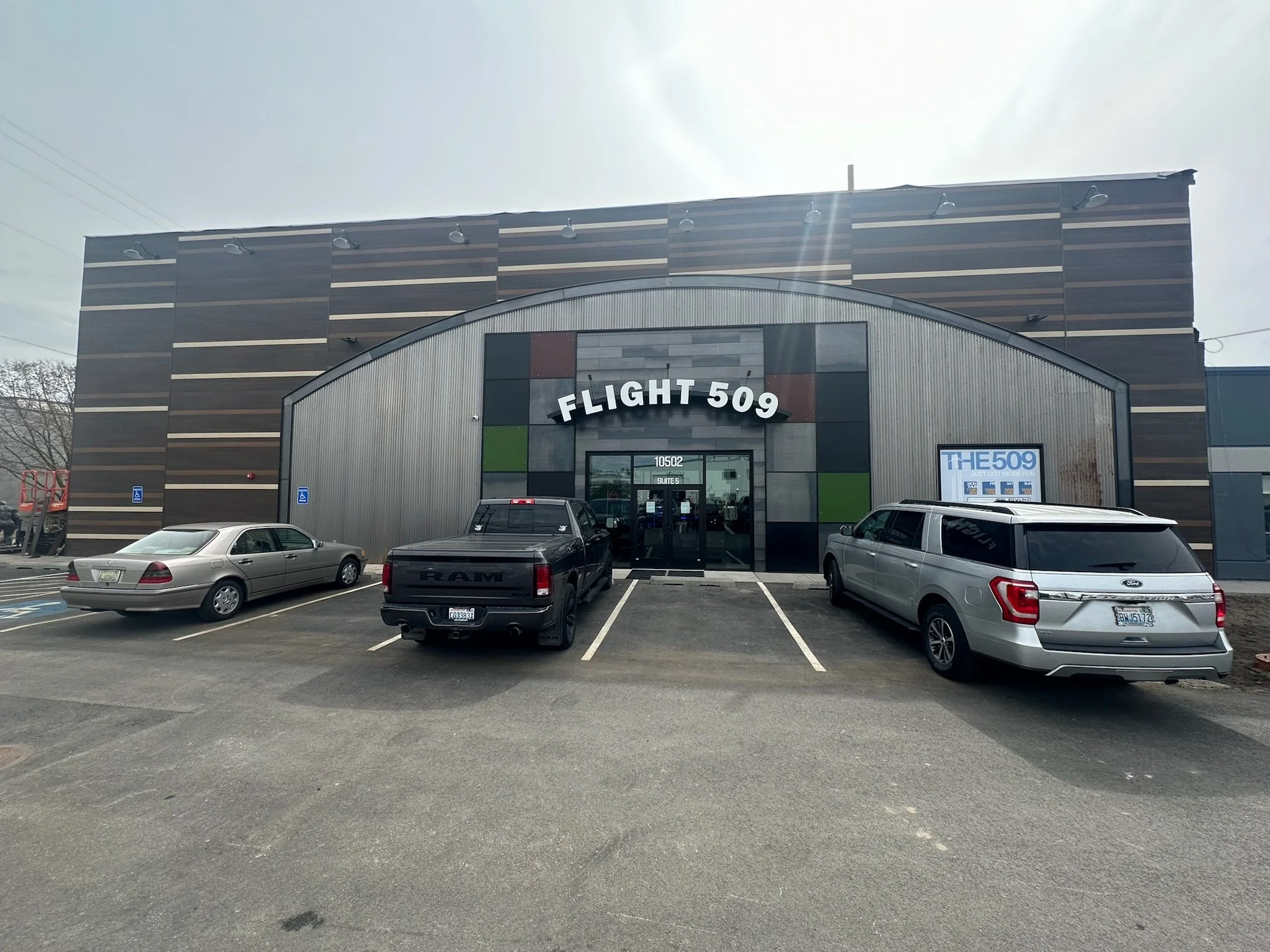 Exterior view of a building named Flight 509 with a parking lot in front containing three cars. The building has a modern design with a curved metal front and dark wood paneling on the upper part. There is a sign on the right side that reads 'The 509