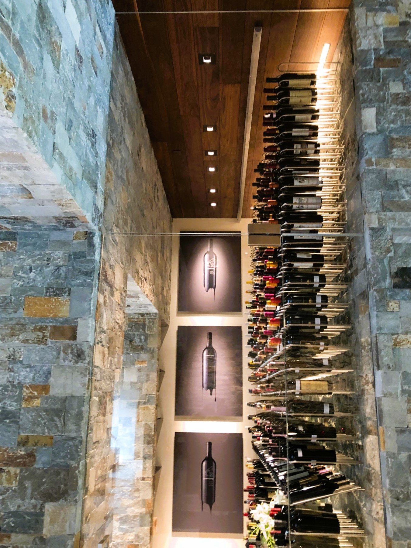 Interior of a wine cellar with stone walls, a wooden ceiling, and a large glass display of wine bottles on a metal rack. There are three black-and-white photographs of wine bottles on the wall.