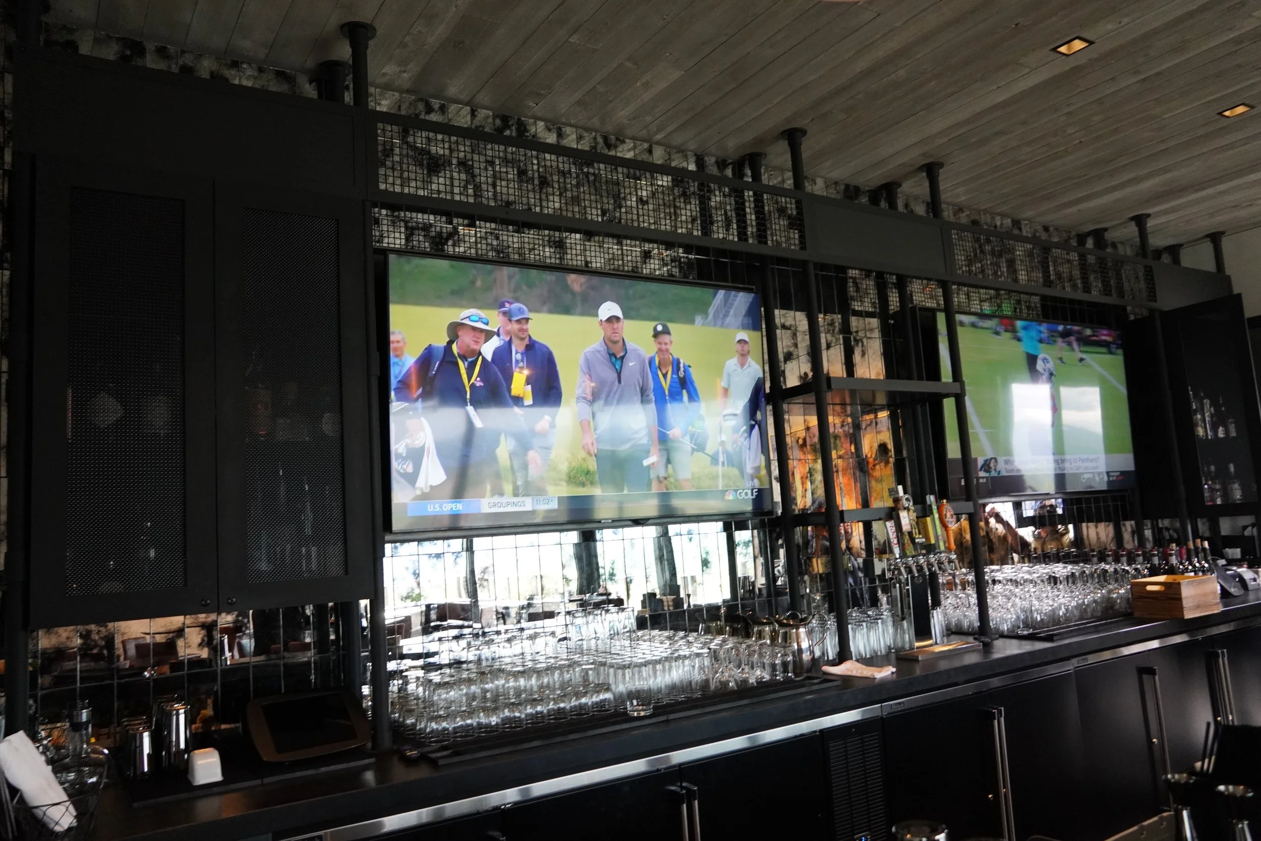 Two flat-screen TVs showing golf tournament scenes, with glasses and bar items on the counter below in a modern bar or restaurant setting.