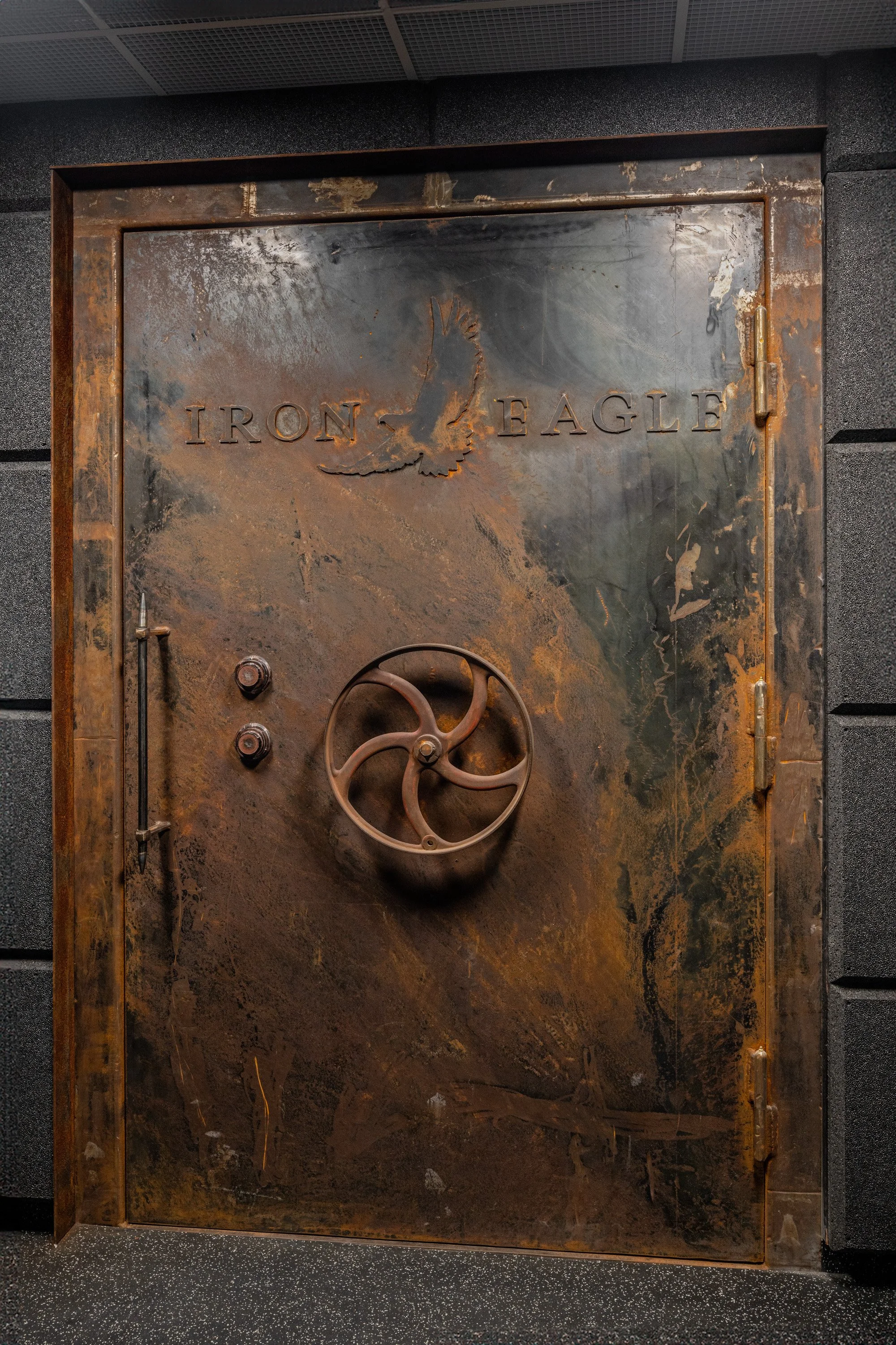 A large, rusty metal vault door with the words 'Iron Eagle' and an eagle engraving. The door features a wheel handle and a latch, set within a dark, textured wall.