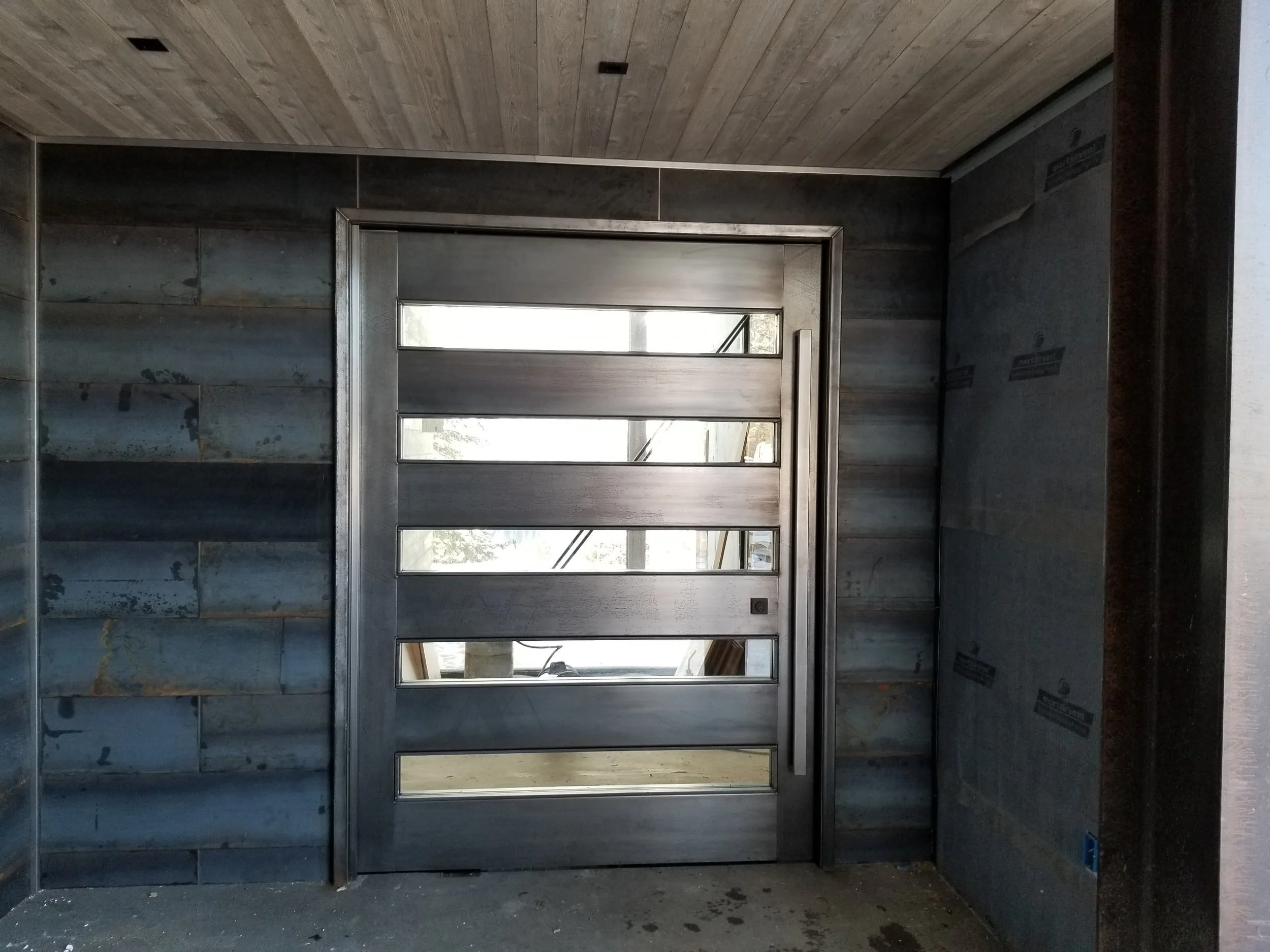 Modern front entrance door with horizontal glass panels, framed by metal, set within a gray wood-paneled wall under a wood ceiling.