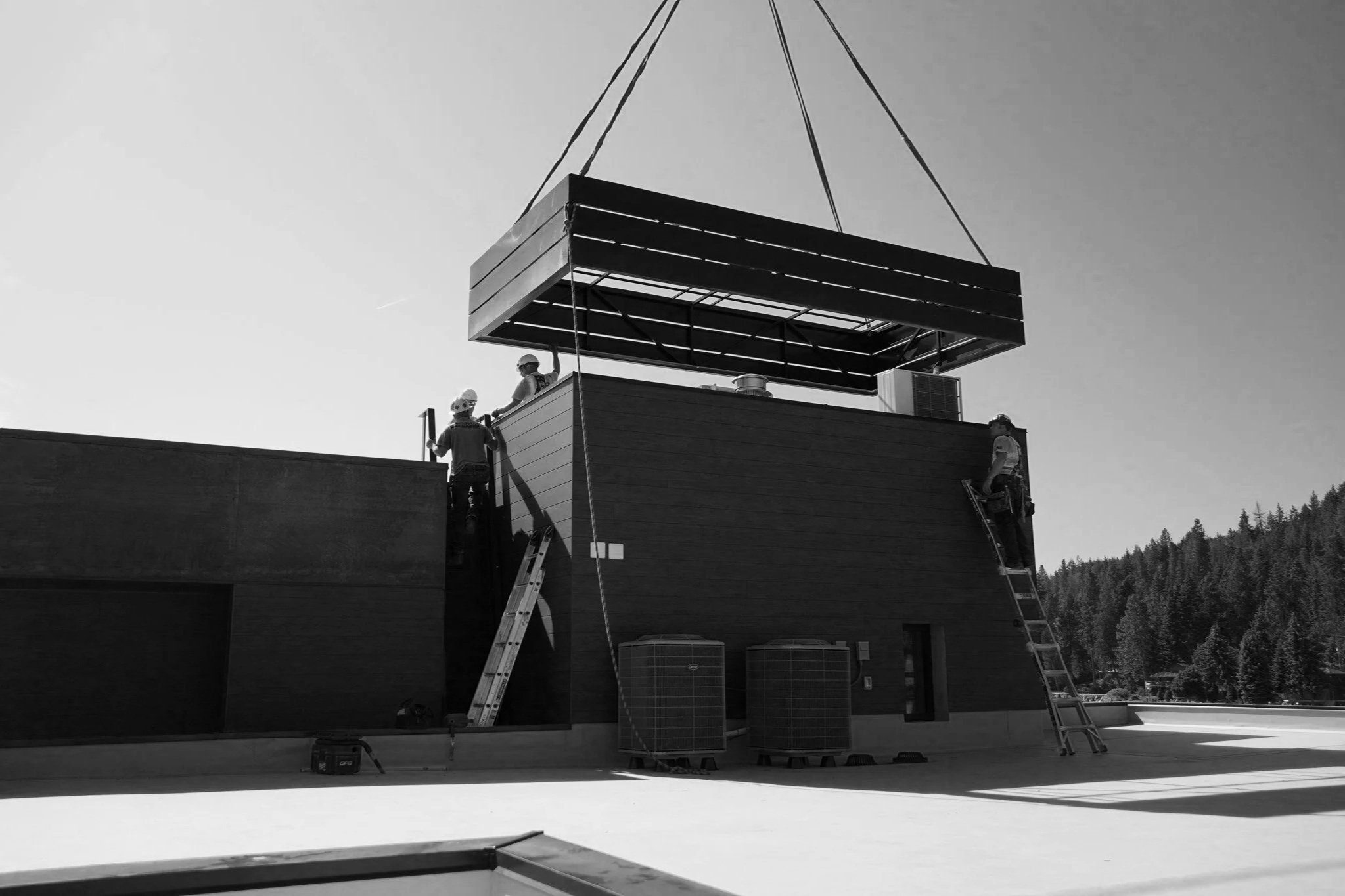 Construction workers installing a large, overhanging structure on a building, with ladders and air conditioning units nearby, in a scenic outdoor setting.