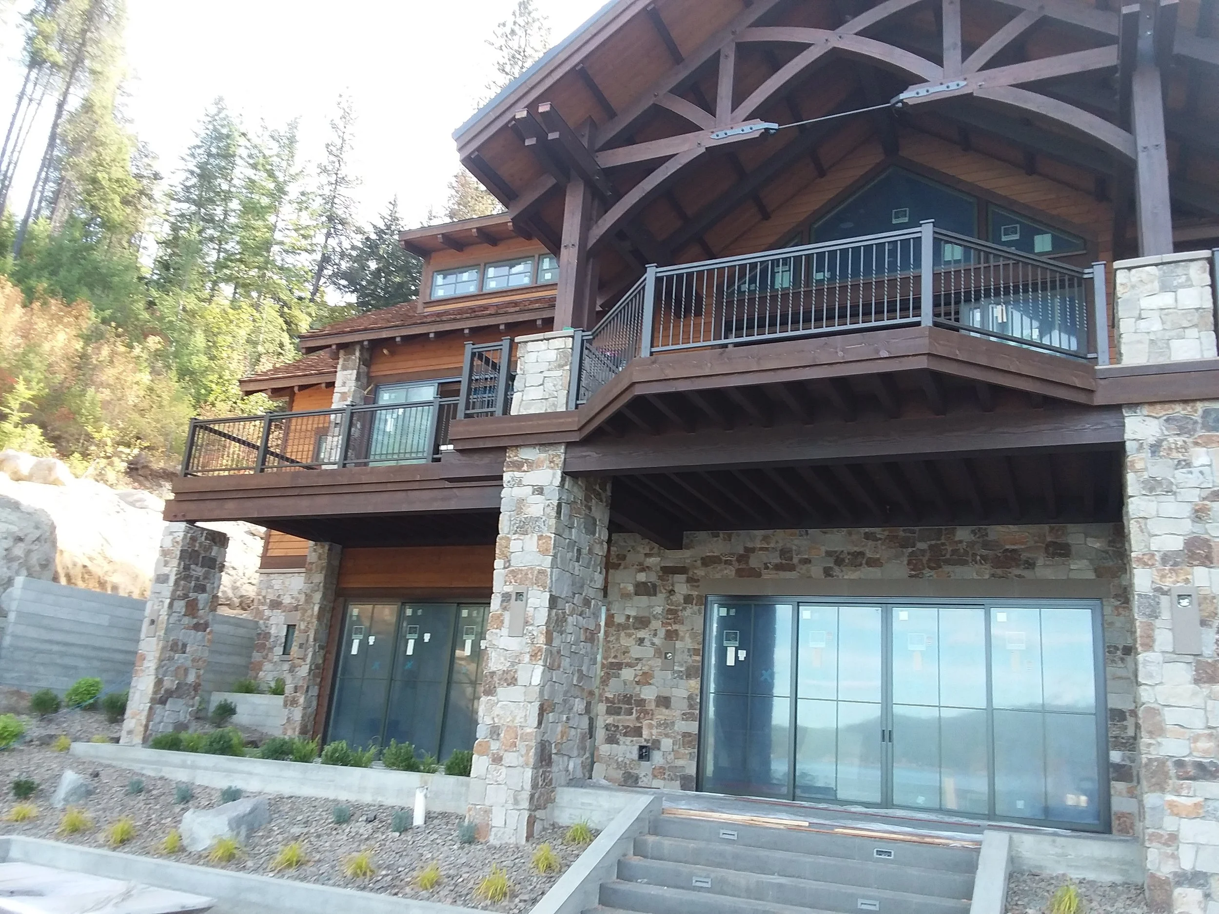 A multi-story house built with stone and wood, featuring large glass sliding doors, a spacious wooden balcony with black railing, and exterior landscaping with small plants and rocks.