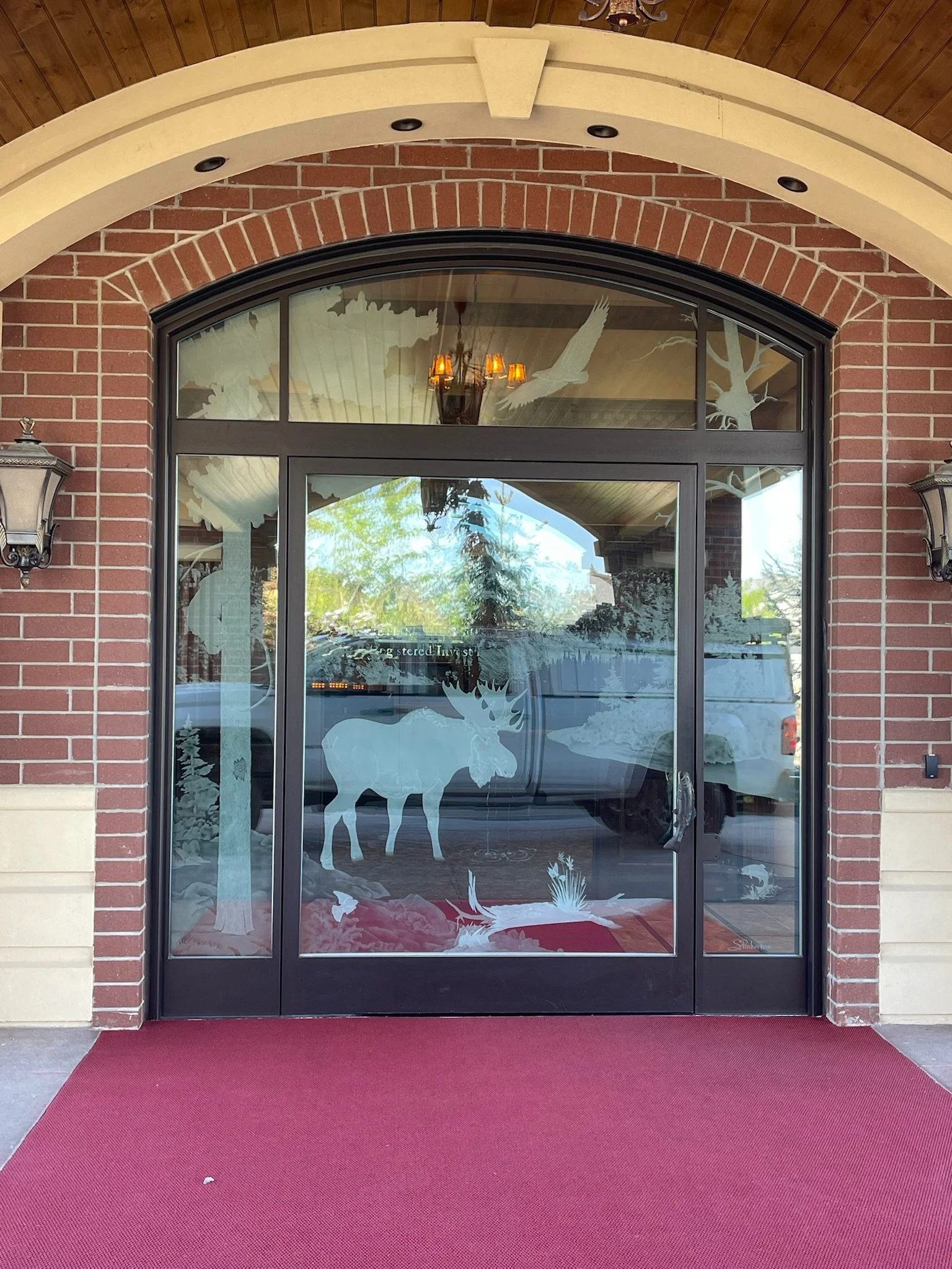 Glass door with winter-themed decals of a moose, trees, and birds, framed by brick walls and a red carpet entry.