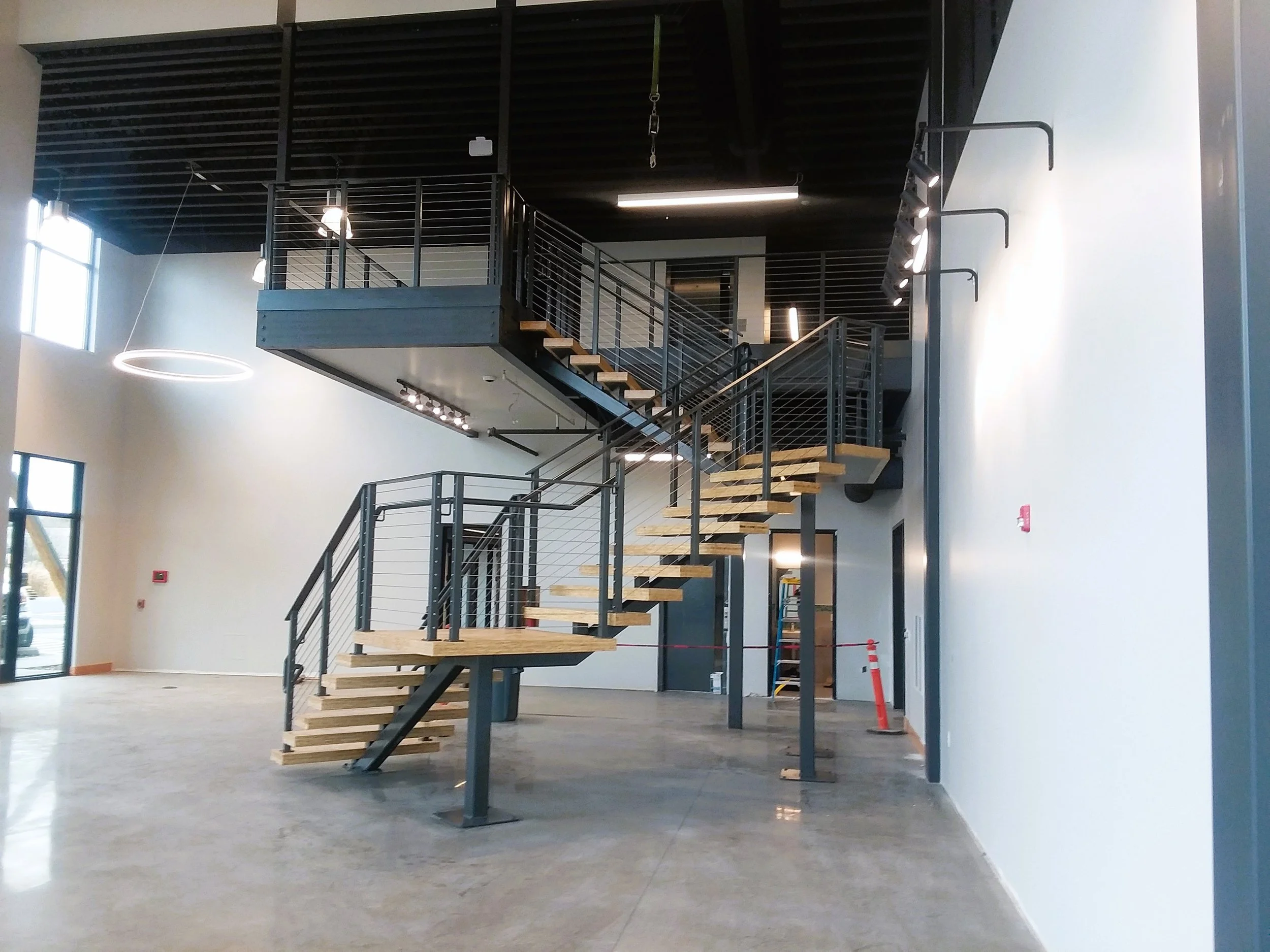 Modern interior with a wooden staircase and black metal railings leading to an upper level, large windows on the left, and a high ceiling with industrial lighting.