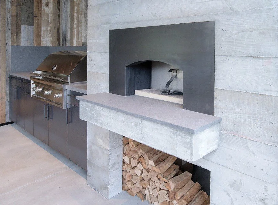 A modern outdoor kitchen with a built-in grill, a concrete fireplace, and firewood stored underneath.