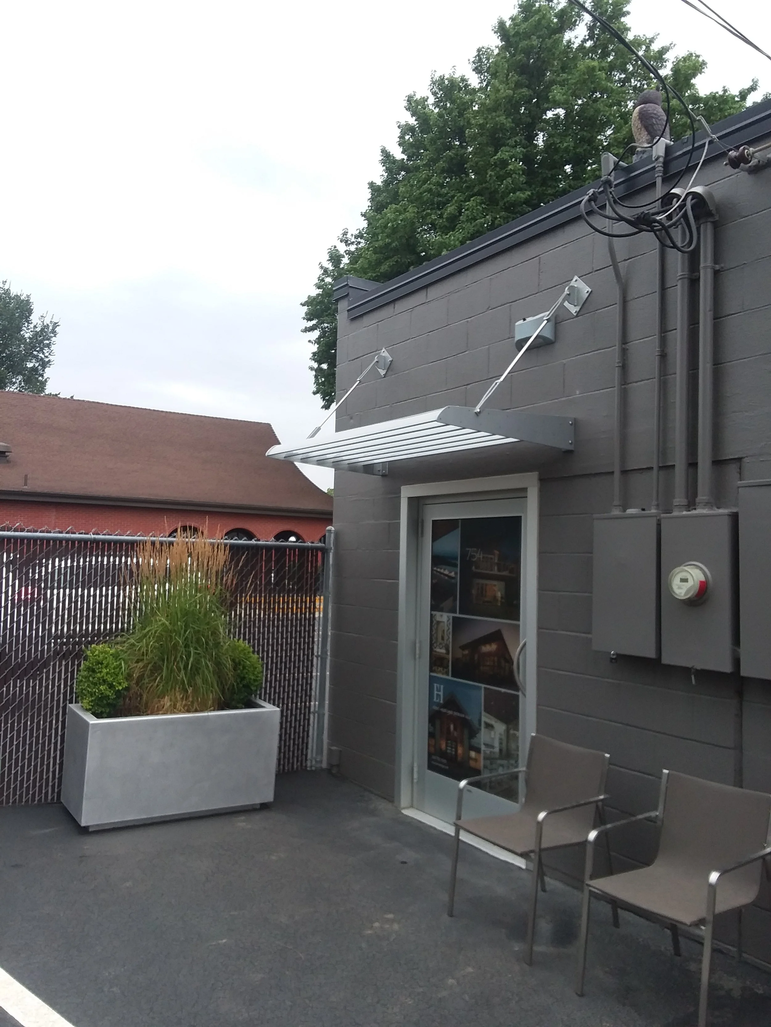 Outdoor patio area with two chairs, a potted plant, a glass door with posters, a gray building wall, and a metal awning above the door.