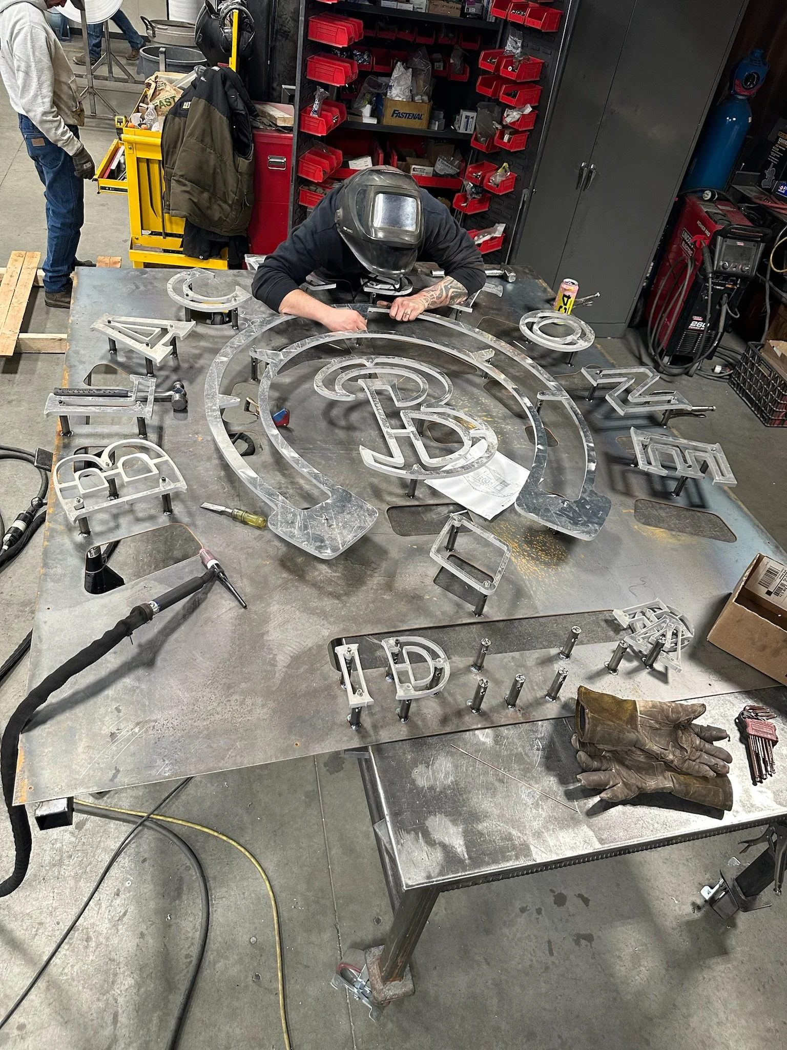 A person wearing a welding helmet working on a large metal sign that spells 'REBEL'. The sign is made of metal letters and logo pieces on a metal worktable. There are welding tools, gloves, and various tools around the workspace.