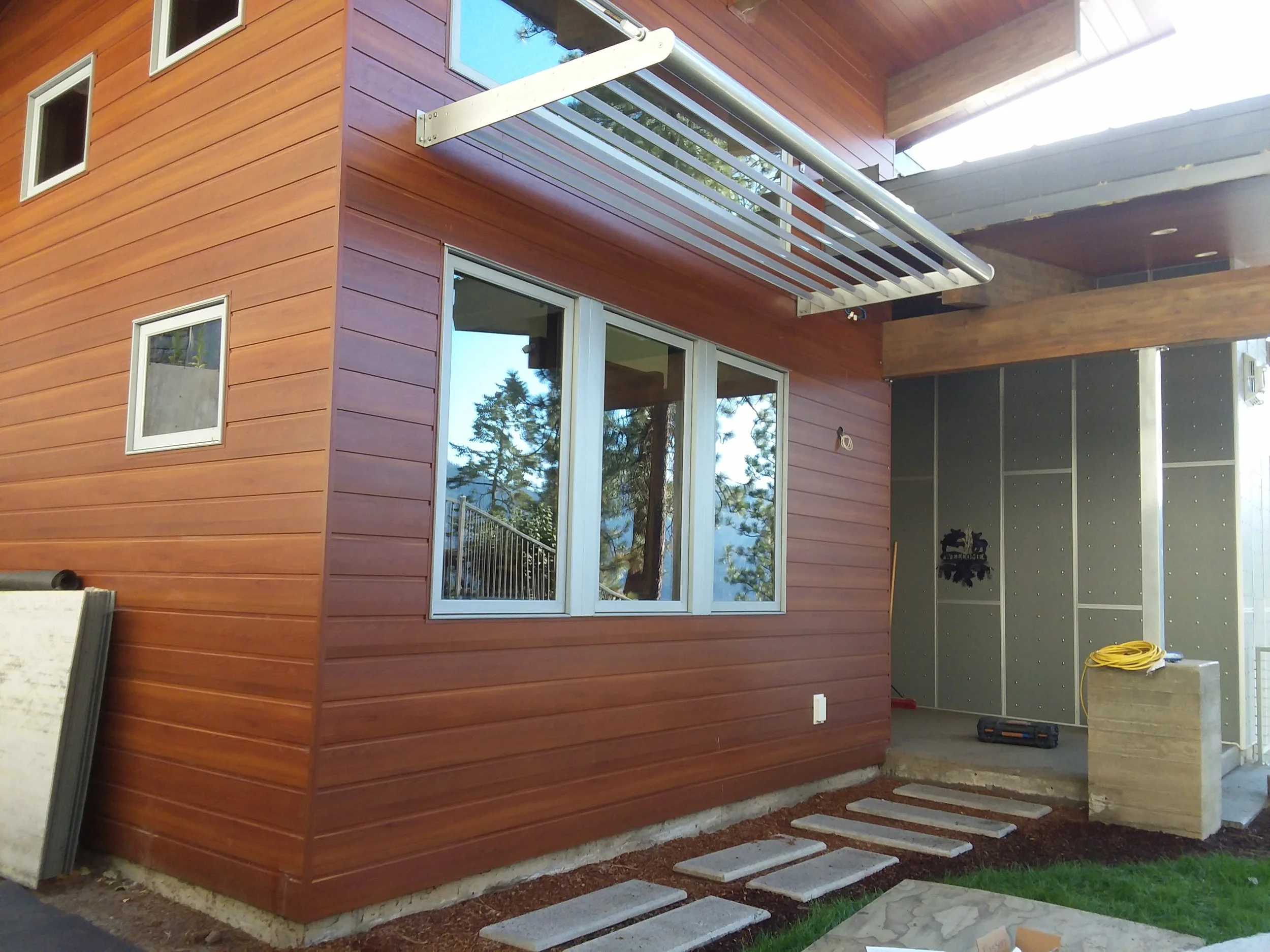 Construction site of a modern house with wooden siding, large windows, and a small patio area with stepping stones and construction materials.