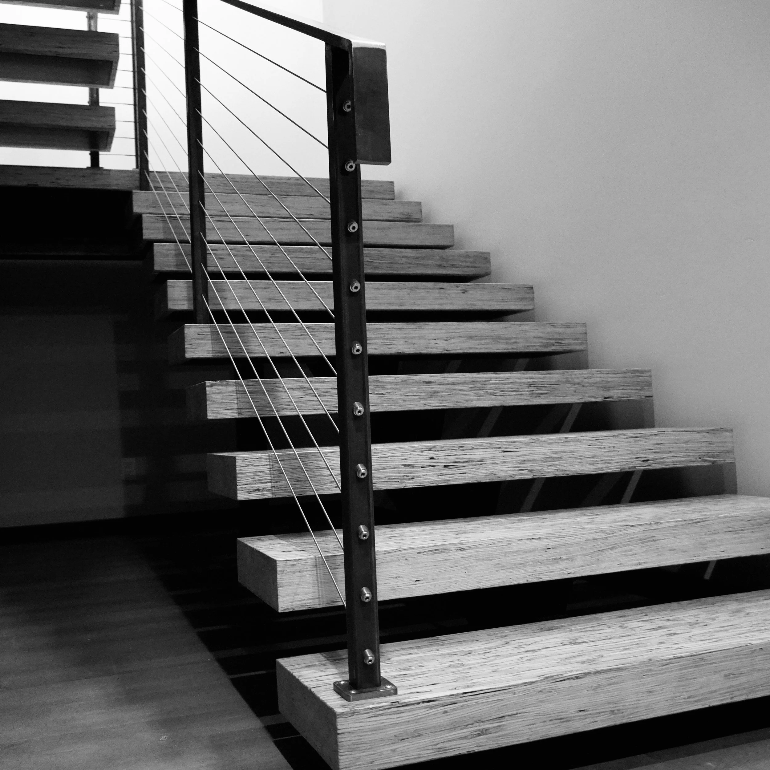 Black and white photo of wooden staircase with metal railing and wires, ascending along the wall.