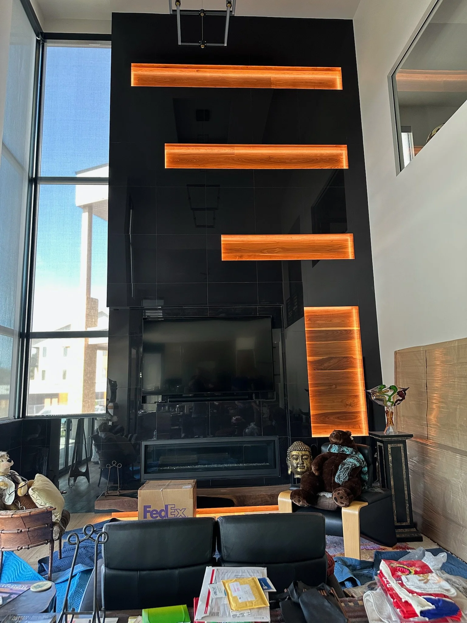Living room featuring a large black wall with wooden illuminated shelves and a centered flat-screen TV, surrounded by unpacked items and decorations.