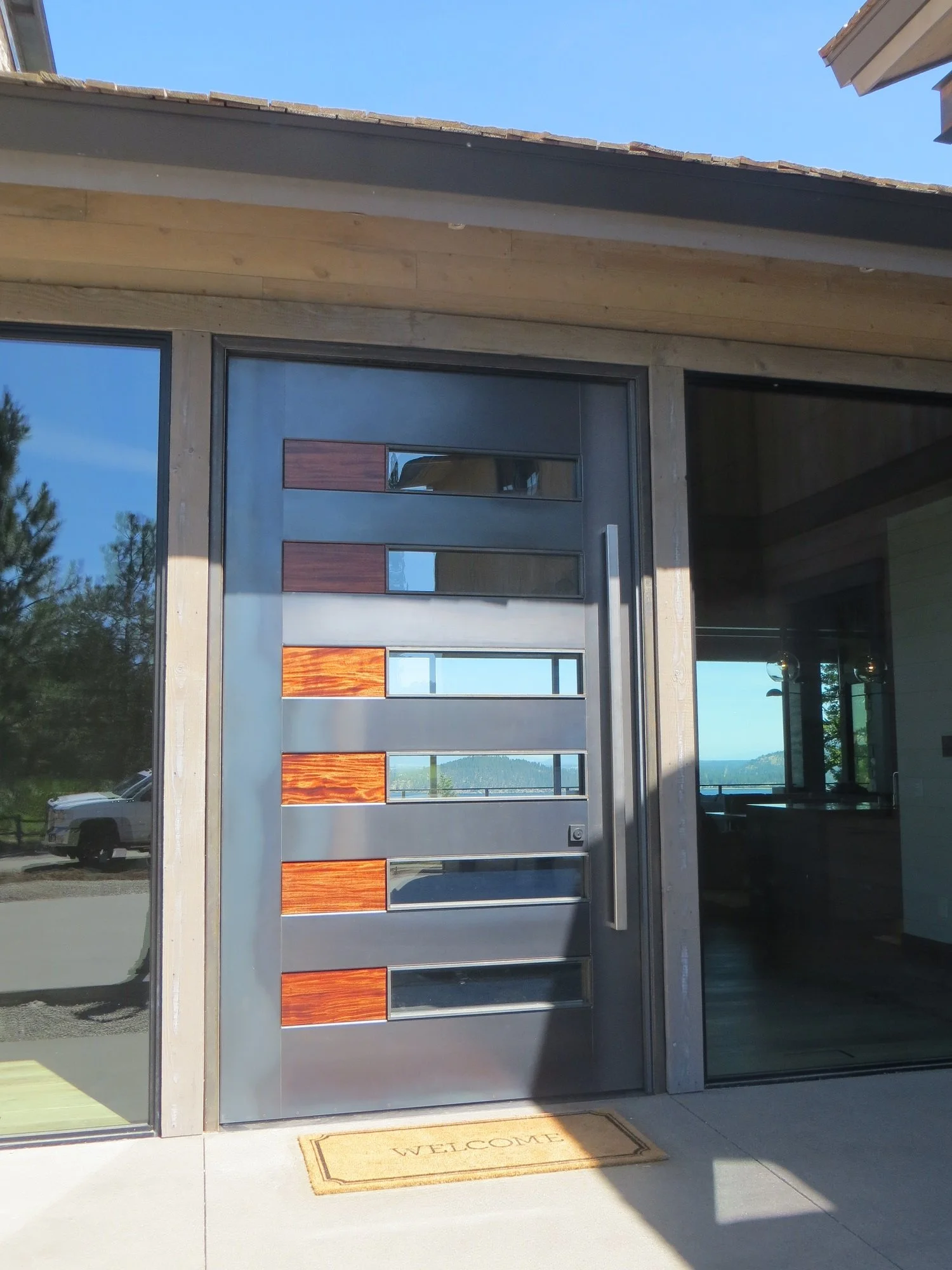 Modern front door with horizontal metal and wooden panels, a long vertical handle, and a welcome mat outside.