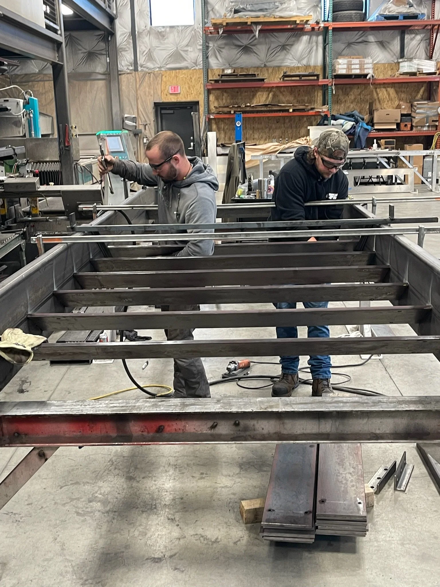 Two men work on a metal structure in a workshop. One man uses a welding machine, and the other is looking down, possibly inspecting parts of the structure.