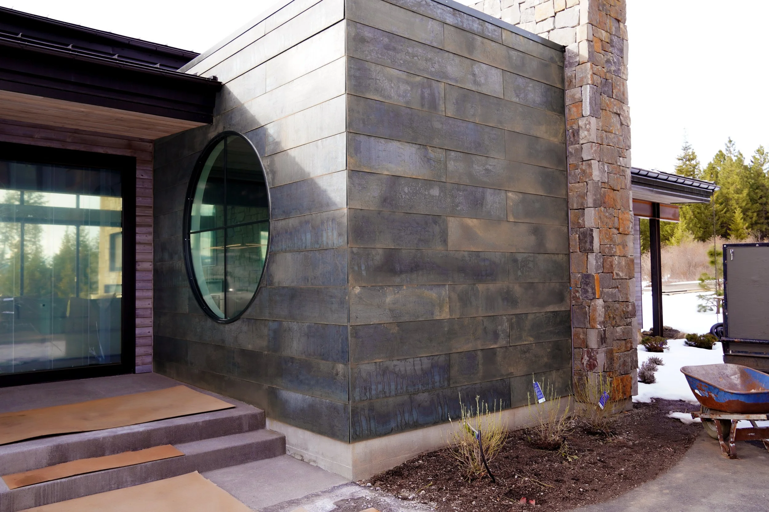 Exterior view of a modern house with a stone and metal facade, featuring a round window on the metal wall, steps leading to a sliding glass door, and a landscaped yard with plants, snow, and a wheelbarrow.