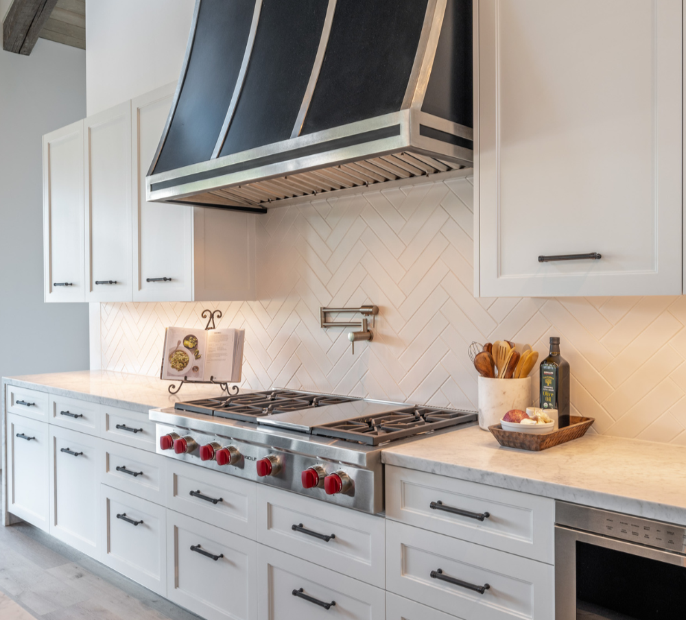 White kitchen cabinets with black handles and a marble countertop, a stainless steel stove with red knobs under a large stainless steel range hood, a cookbook stand with an open cookbook, a container with wooden cooking utensils, a bottle of olive oil, and a small tray with onions and garlic.