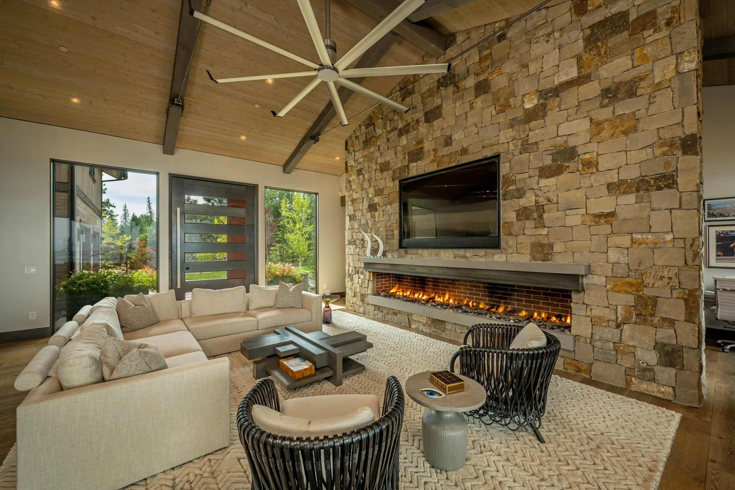 Living room with beige sectional sofa, two black woven chairs, and a small round side table, with a large stone fireplace, a wall-mounted TV, and a wooden ceiling with a modern ceiling fan, near glass doors leading outside.