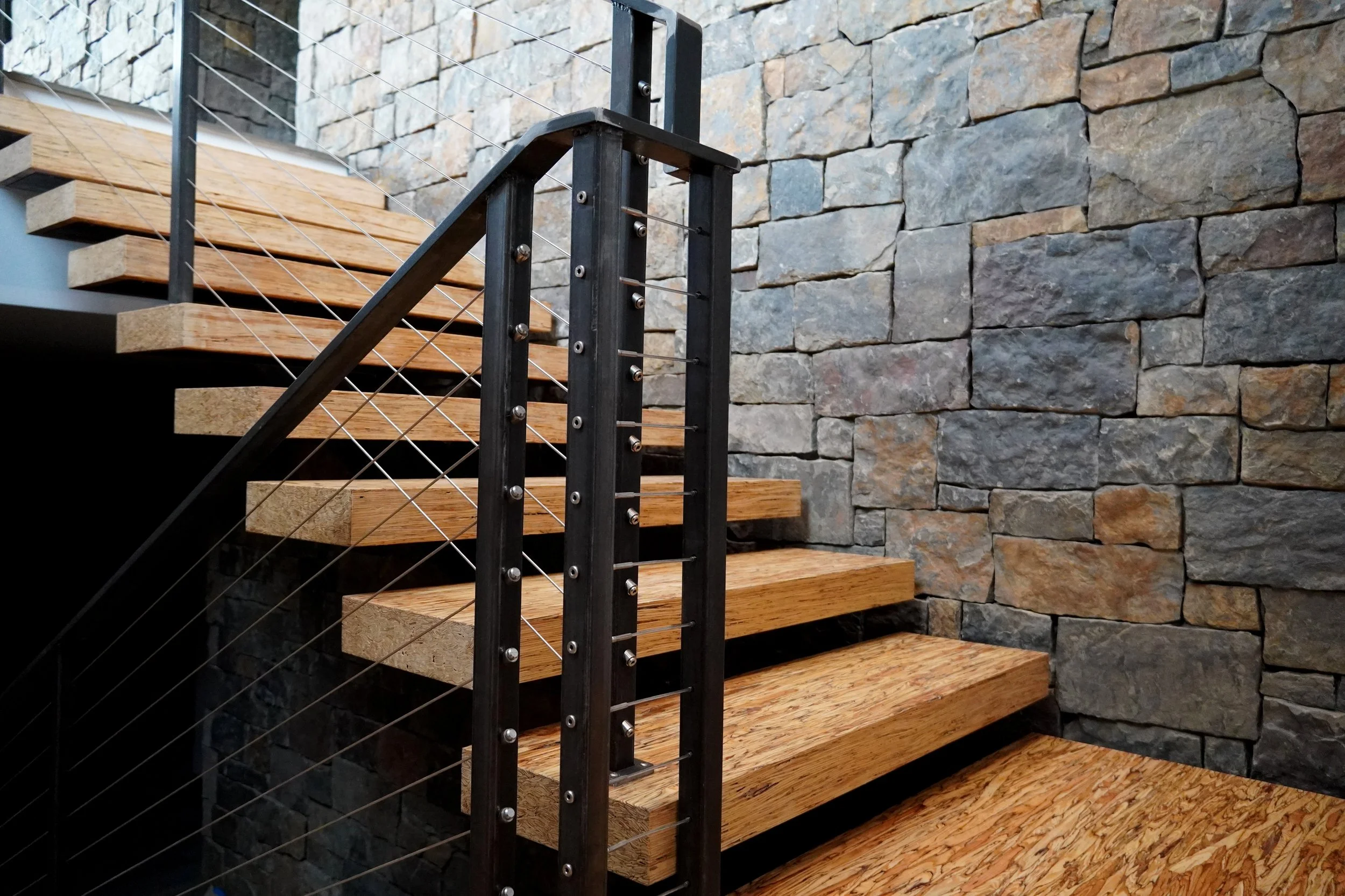 Interior staircase with wooden steps, a black metal railing with wire cables, and a stone wall background.