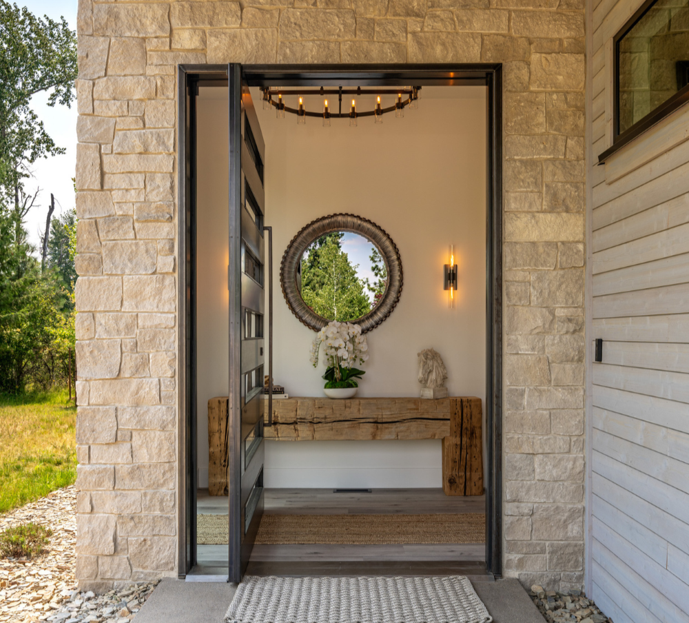 View into a modern entryway with a rustic wooden console table adorned with a white orchid, a stone sculpture, and a circular mirror on a white wall. A black sconce light and a chandelier are also visible.