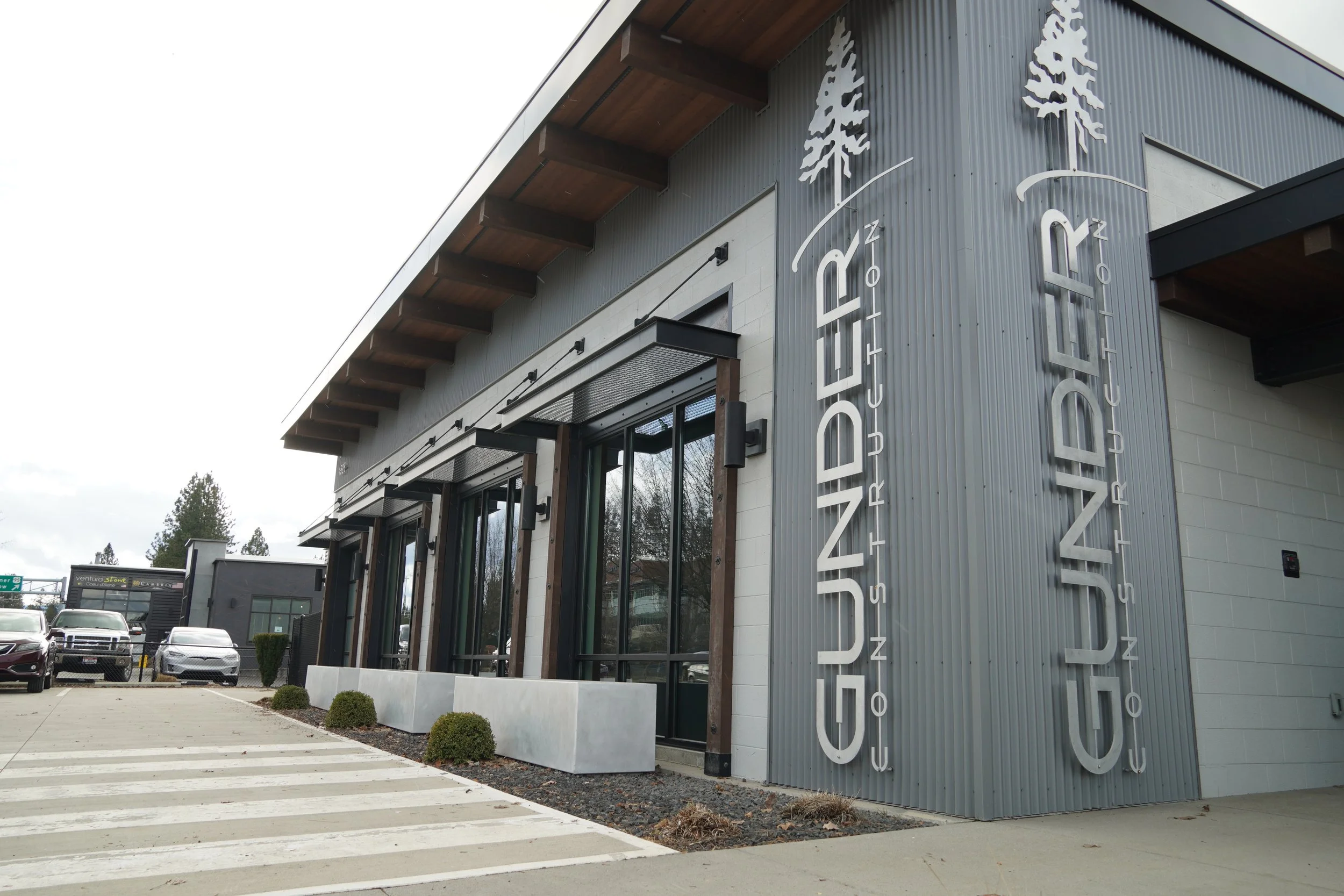 Exterior of a modern building with vertical gray panels and wooden accents, and large vertical signage reading 'MARCH' and 'ZINC.' There are parked cars and a crosswalk in front of the building.