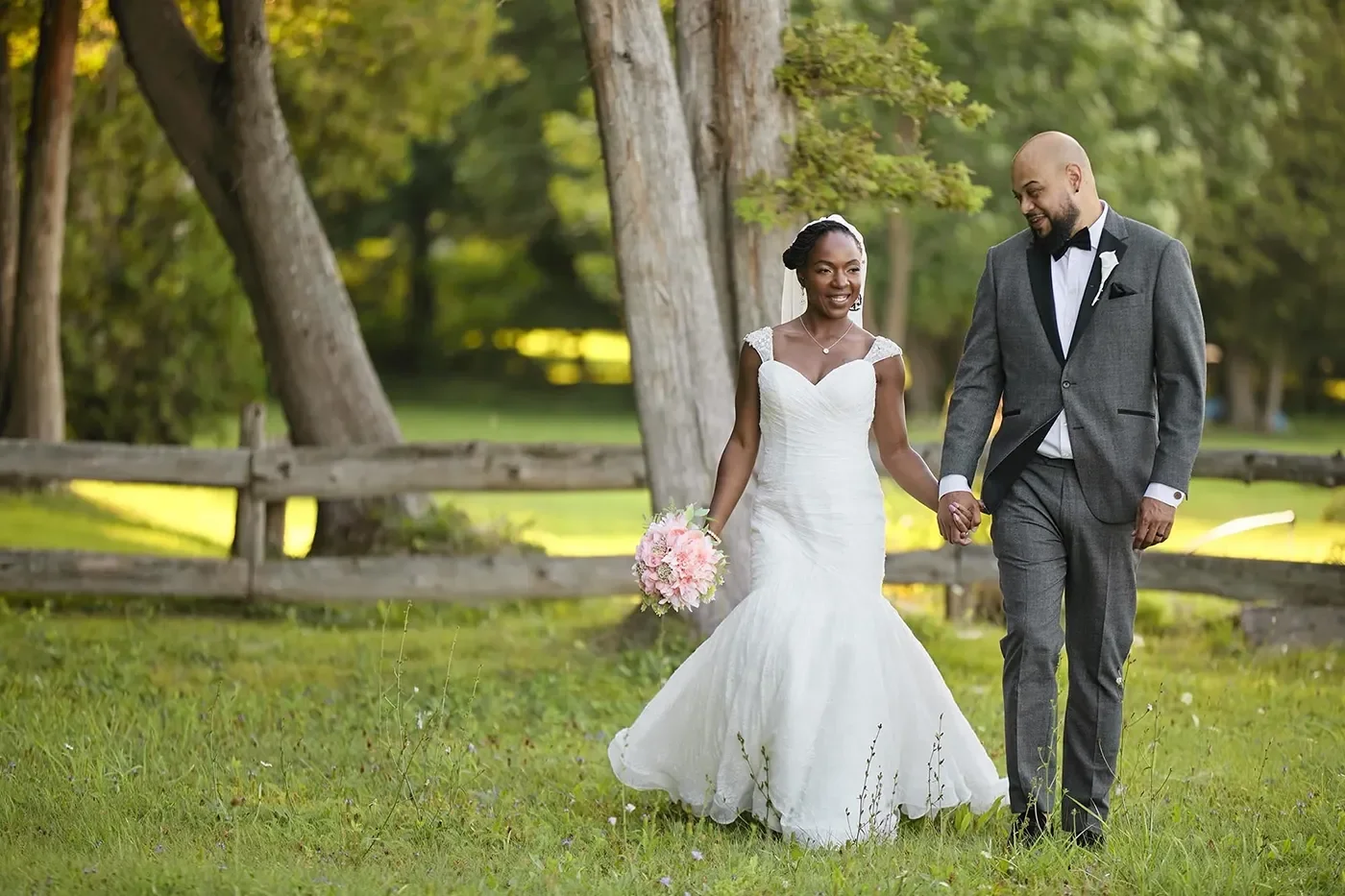 A bride and groom walking together while holding hands in a shaded field on a sunny day.