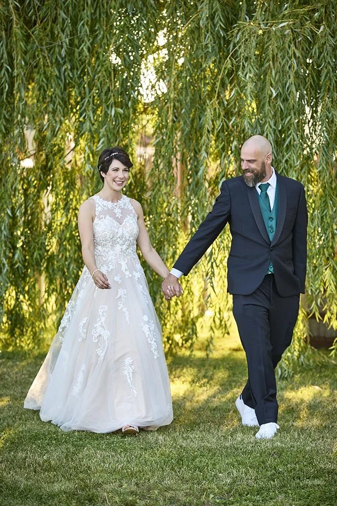 A bride and groom walking outdoors under hanging tree branches, both smiling and holding hands.