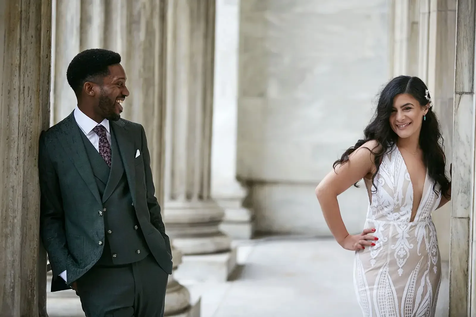 A man and a woman smiling and looking at each other in an elegant corridor with stone columns and arches. The man is wearing a dark suit and the woman is wearing a white dress.
