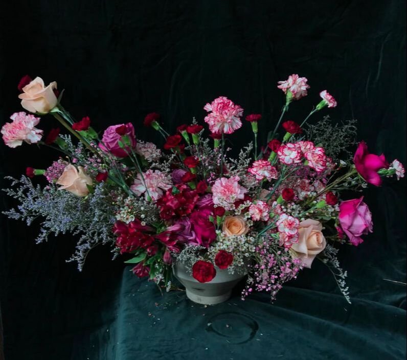 A large floral arrangement in a white vase featuring pink, red, and peach roses, carnations, and other mixed flowers with greenery and delicate filler flowers.