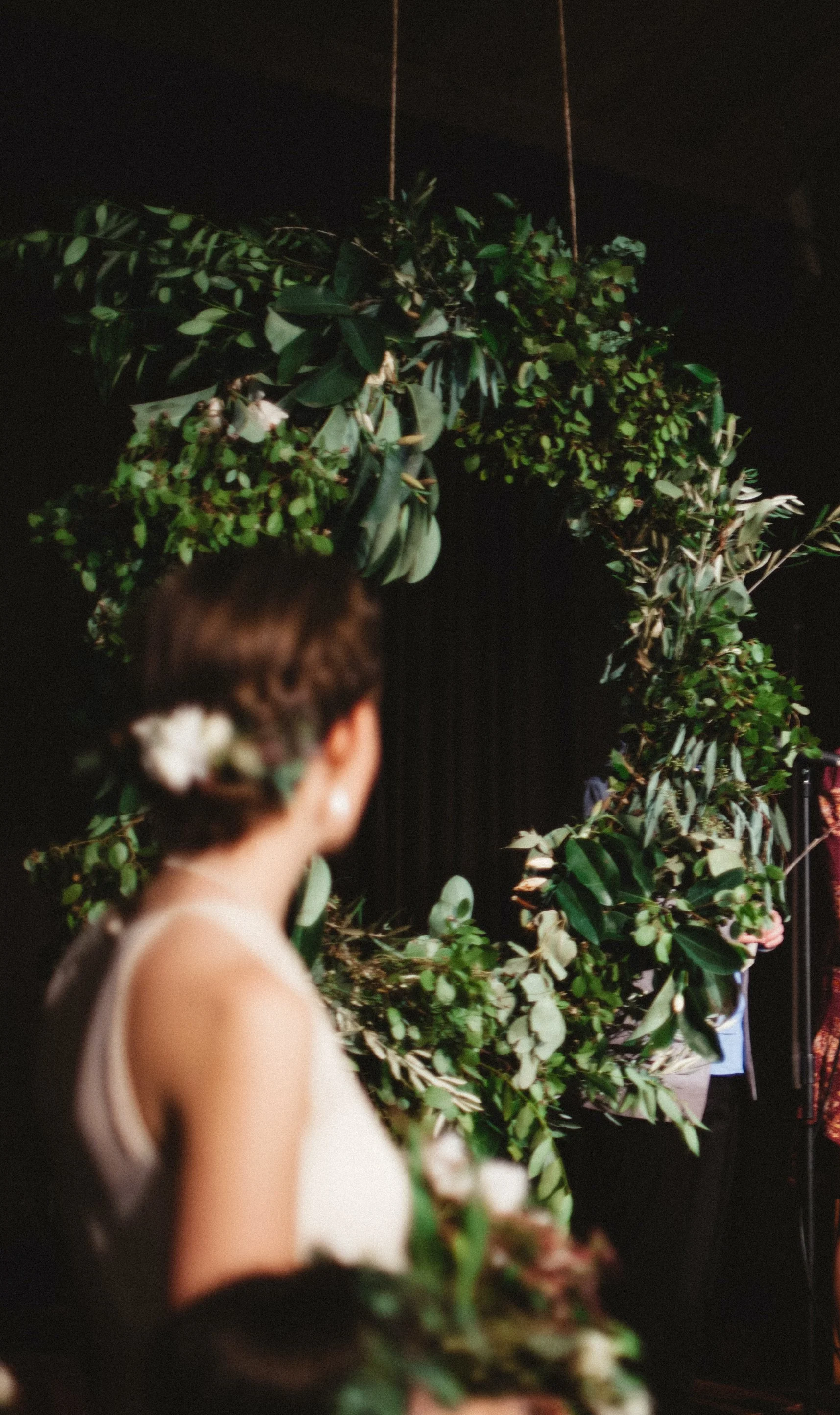 A woman with a flower in her hair standing in front of a large floral arch at what appears to be a wedding or special event.