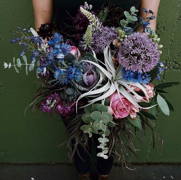 Person holding a large bouquet of mixed flowers including pink roses, purple allium, blue thistle, and various greenery in front of a green wall.