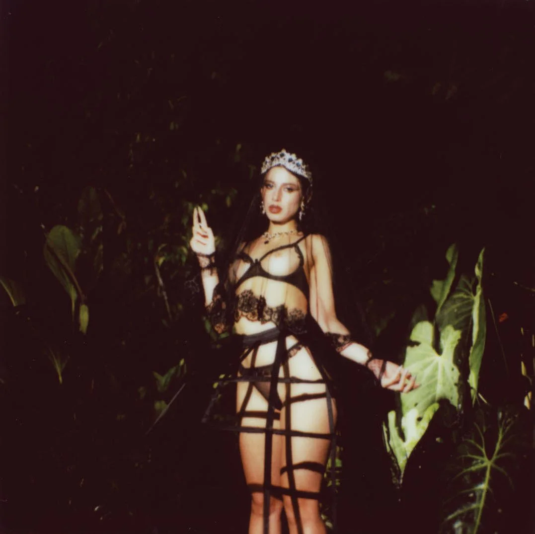 A woman wearing black lace lingerie and a crown, posing in front of large green leaves at night.