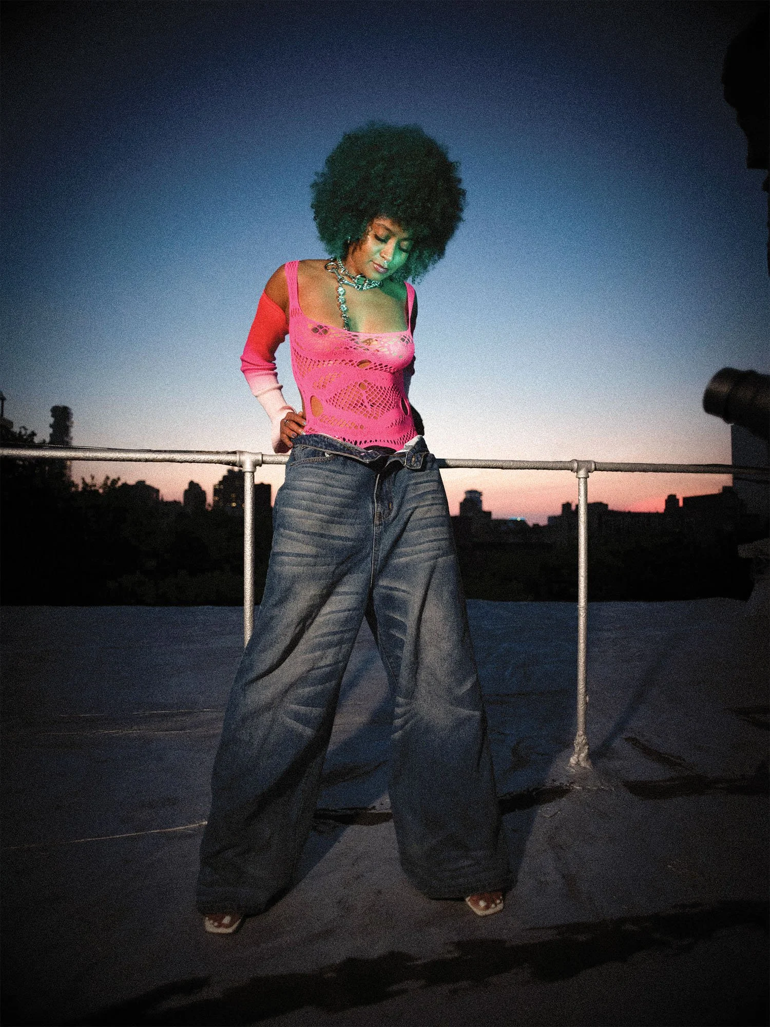 A woman with curly hair wearing a pink crochet top, large silver chain necklace, and wide-leg denim jeans standing on a rooftop during sunset.
