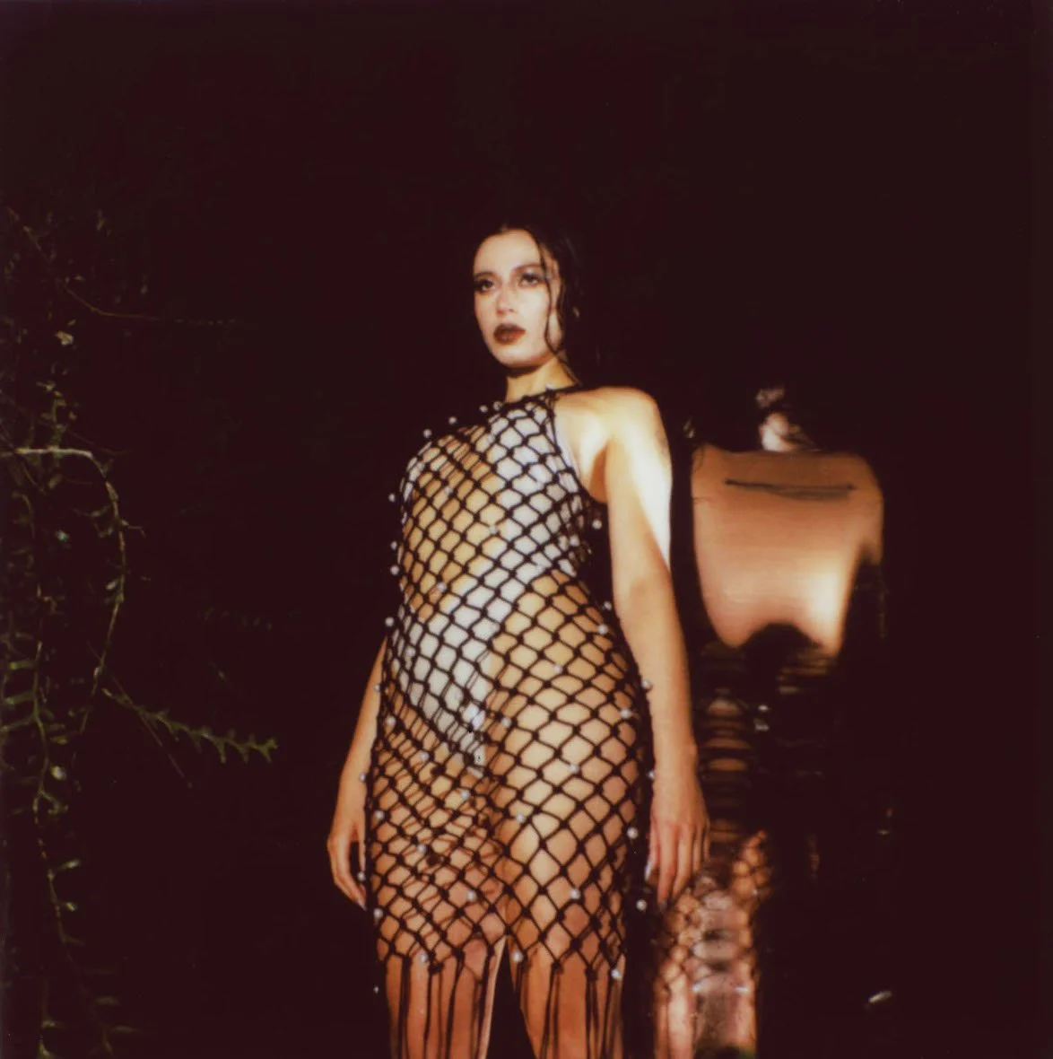 A woman wearing a black fishnet dress with pearl accents standing near a body of water at night.