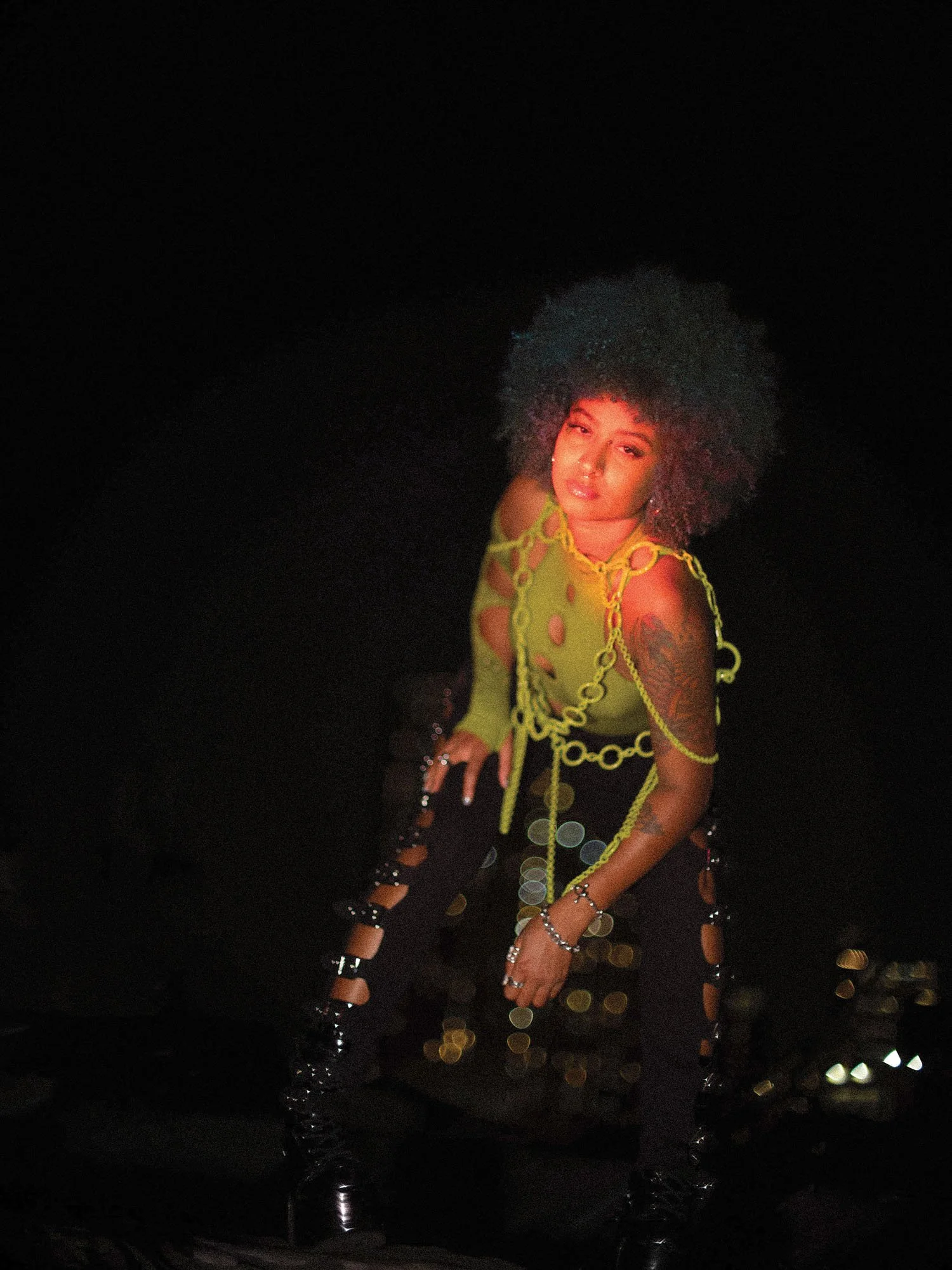 A woman with curly hair wearing a bright yellow chain-link style top and black pants with cutouts, standing in a dark setting with city lights in the background.