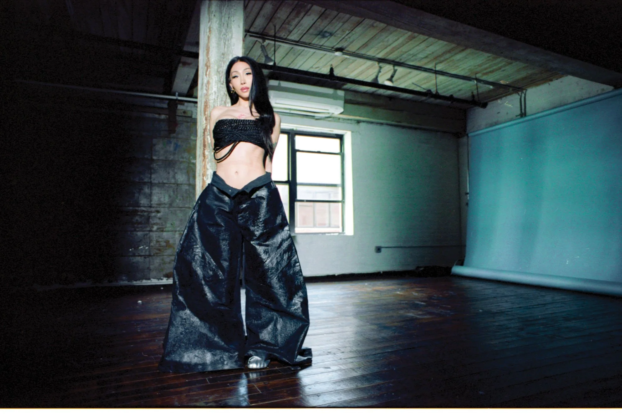 A woman with long black hair and light skin, standing in an industrial-style room with wooden floors, a large window, and exposed ceiling beams. She is wearing a black off-the-shoulder crop top and black wide-leg pants.