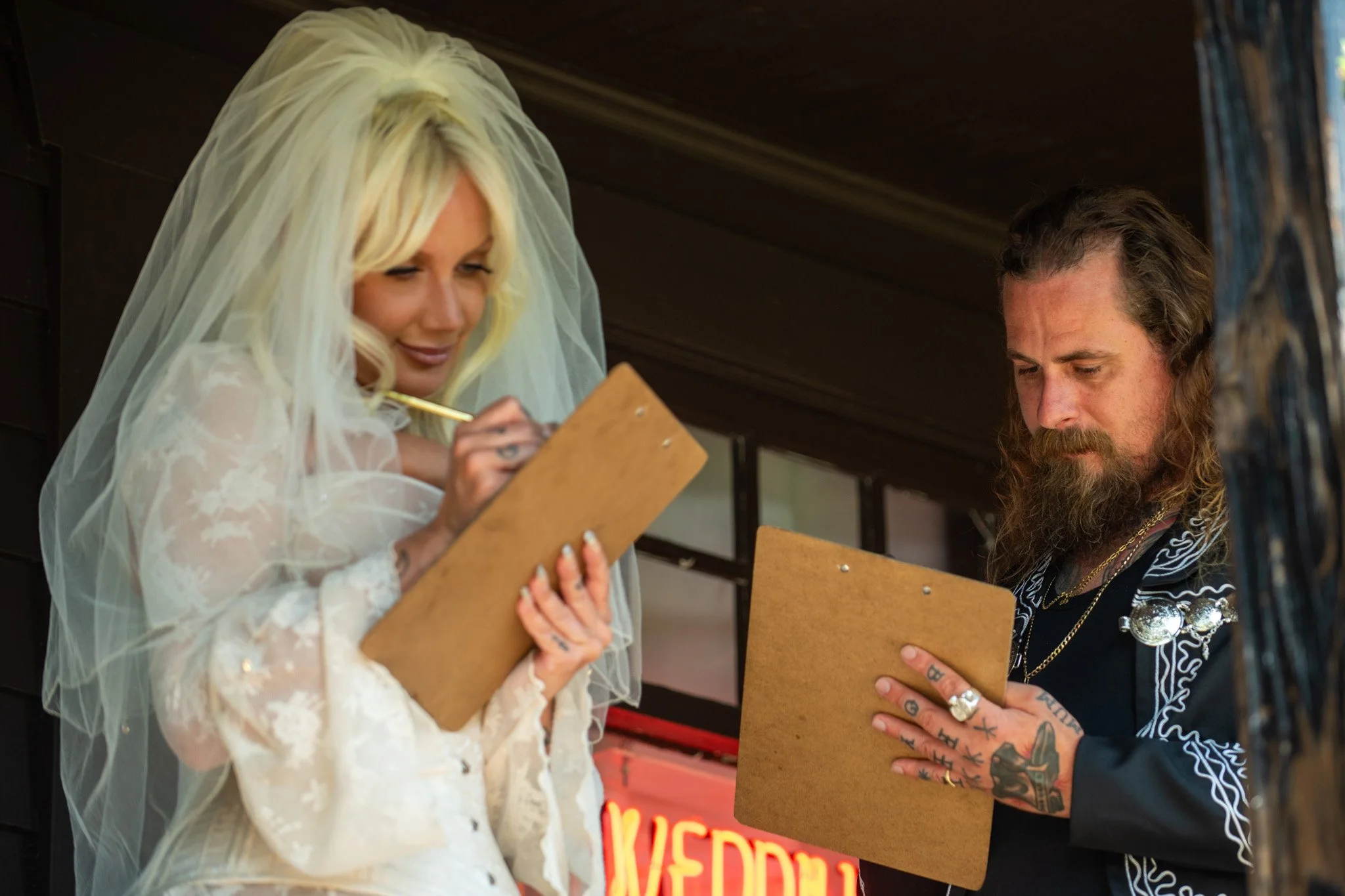 A bride with blonde hair wearing a veil and white lace dress signs a document on a clipboard held by a man with long hair, a beard, tattoos, and wearing a black shirt with white embroidery.