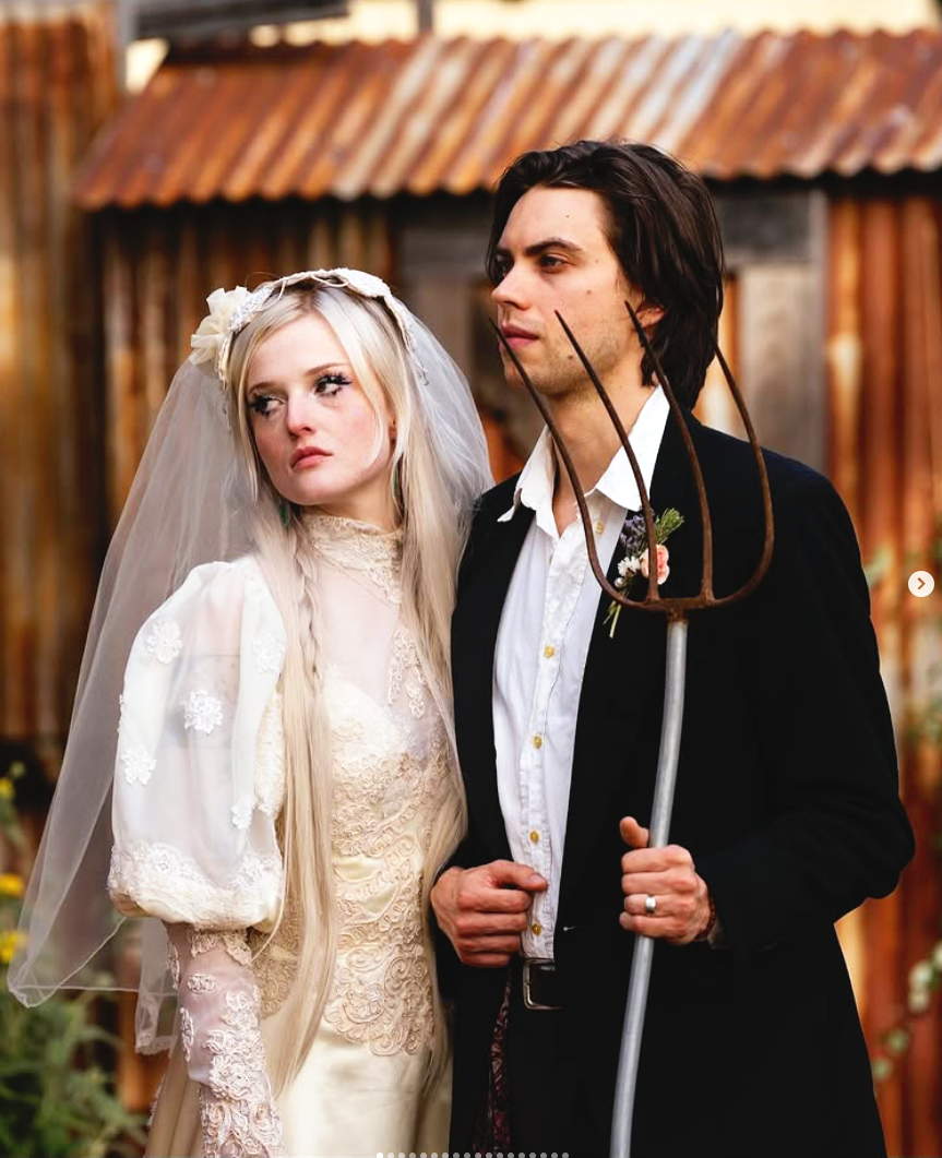 A young woman in vintage wedding dress with lace details and a veil, standing next to a man in a black suit holding a pitchfork, in front of a rustic wooden background.