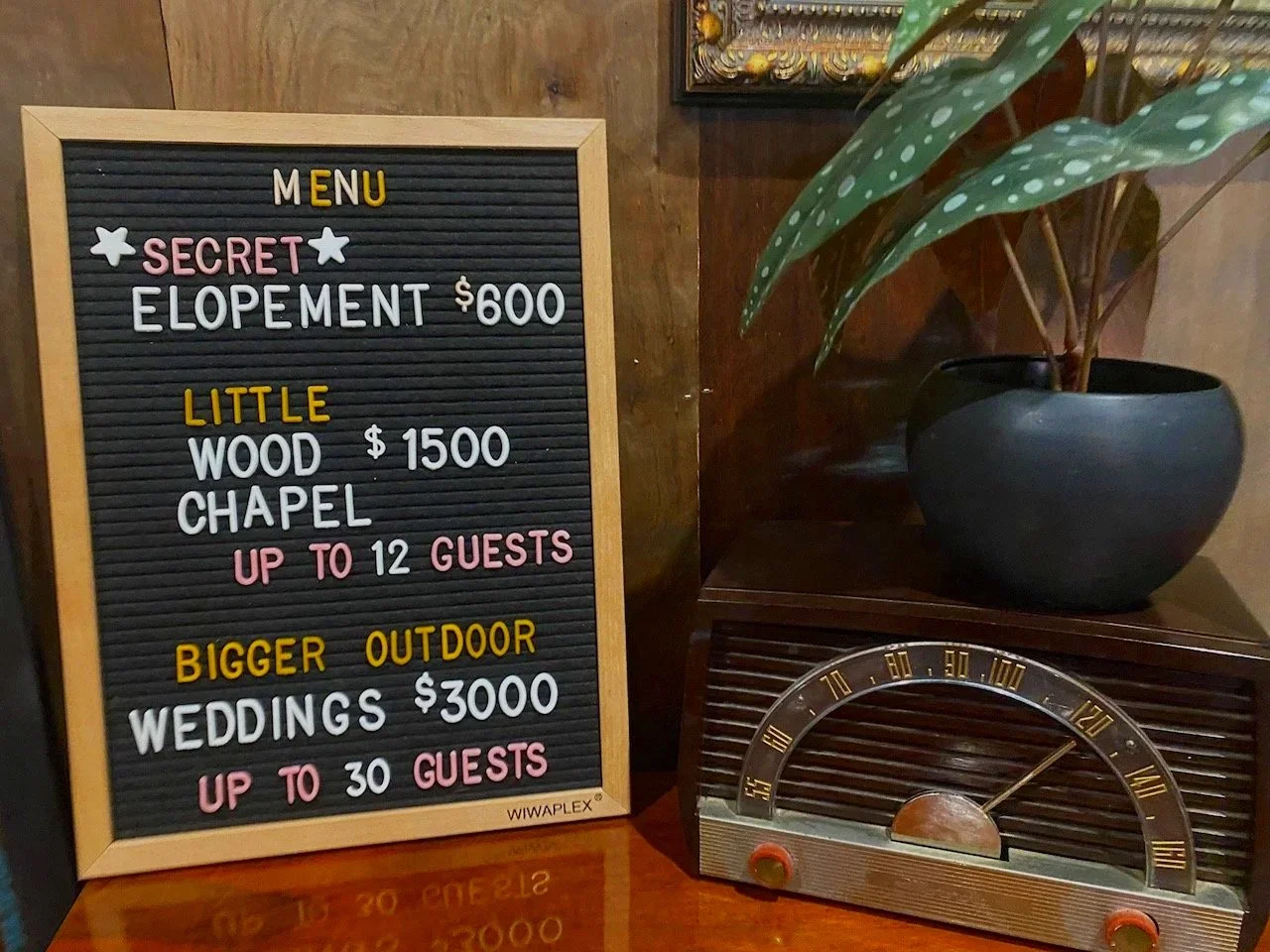 Menu board listing different event packages with prices: Secret Elopement for $600, Little Wood Chapel for $1,500, and Bigger Outdoor Weddings for $3,000, accommodating up to 12 or 30 guests, beside a vintage radio and a potted plant.