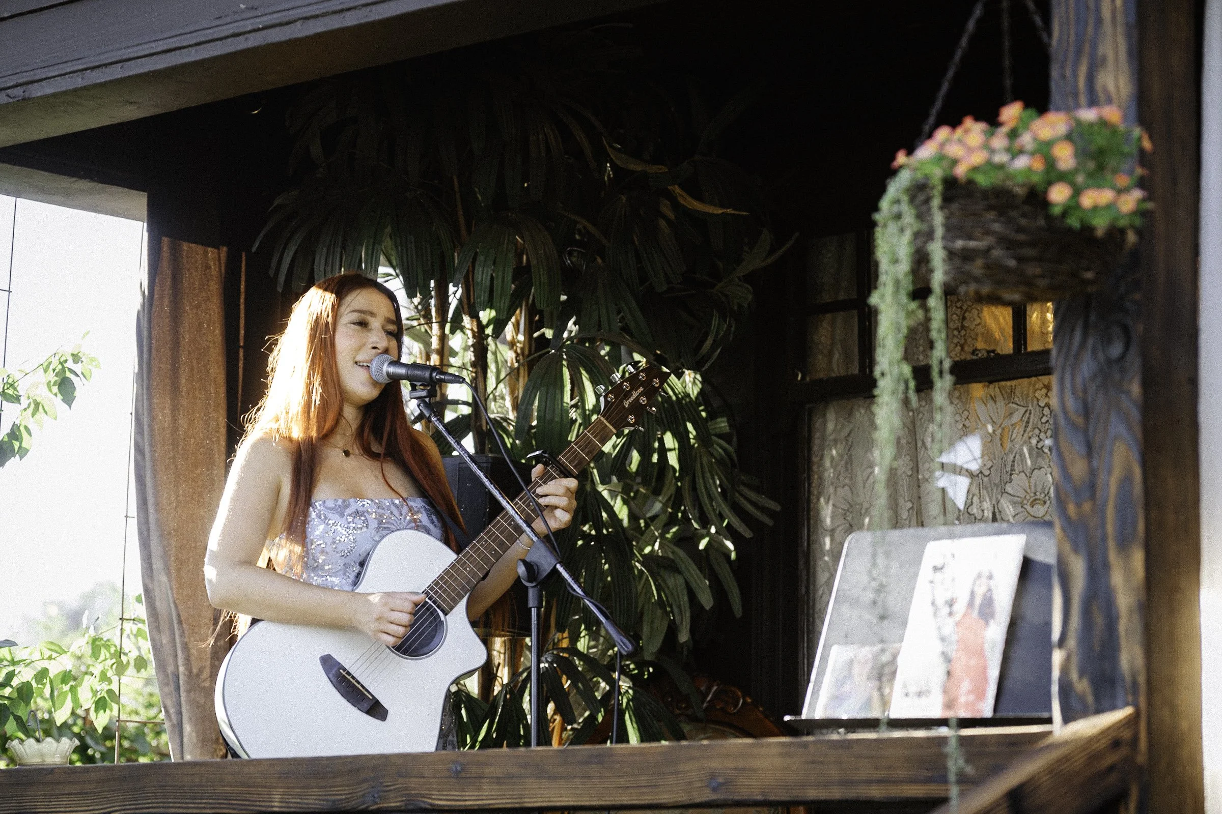 Young woman with long red hair singing and playing acoustic guitar, standing in a cozy room with plants and a window, with a laptop and photos on a stand nearby.
