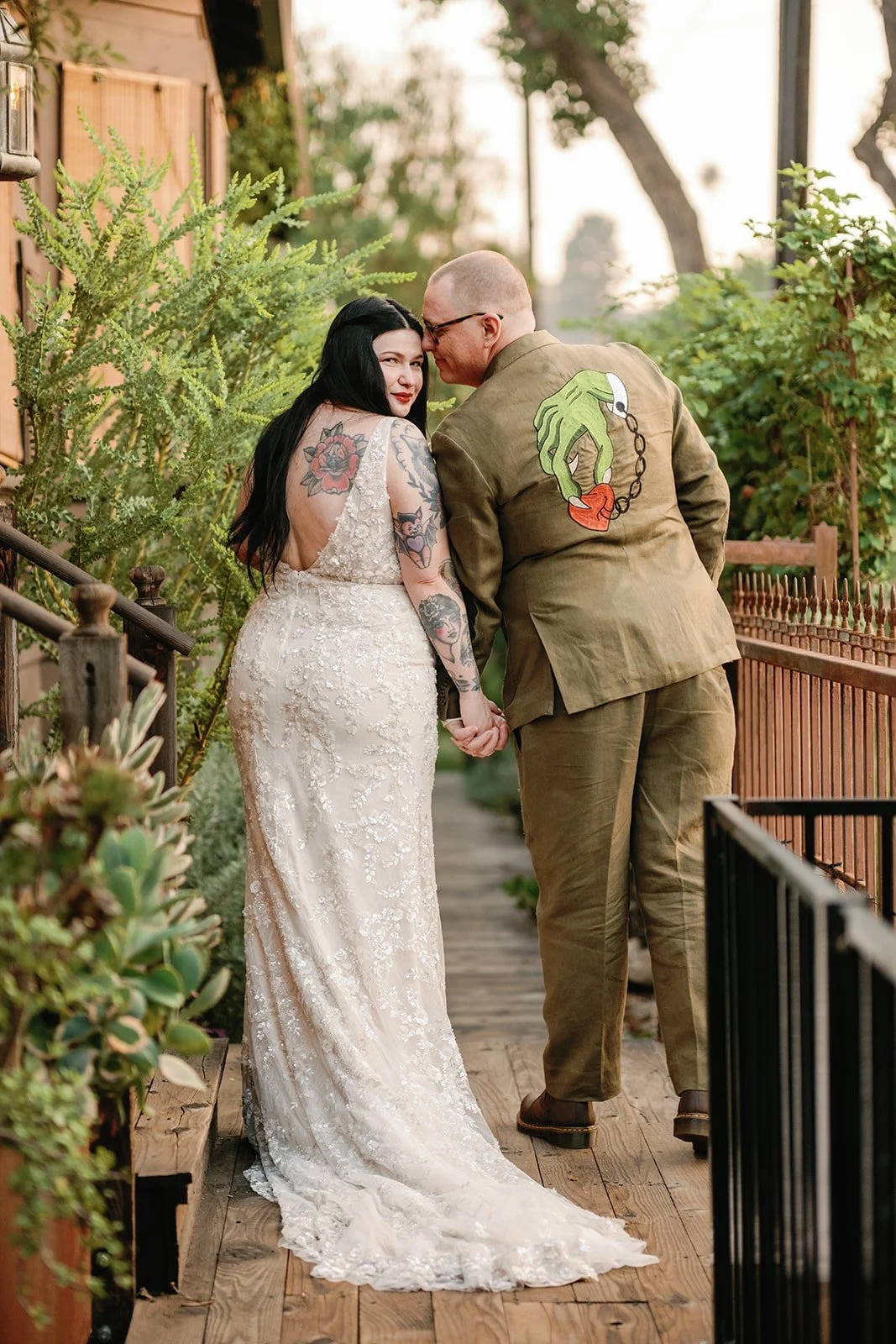 A couple holding hands outdoors on a wooden pathway, the woman with black hair and tattoos in a white lace dress, the man in a green suit with a patch on the back, surrounded by green plants and trees.
