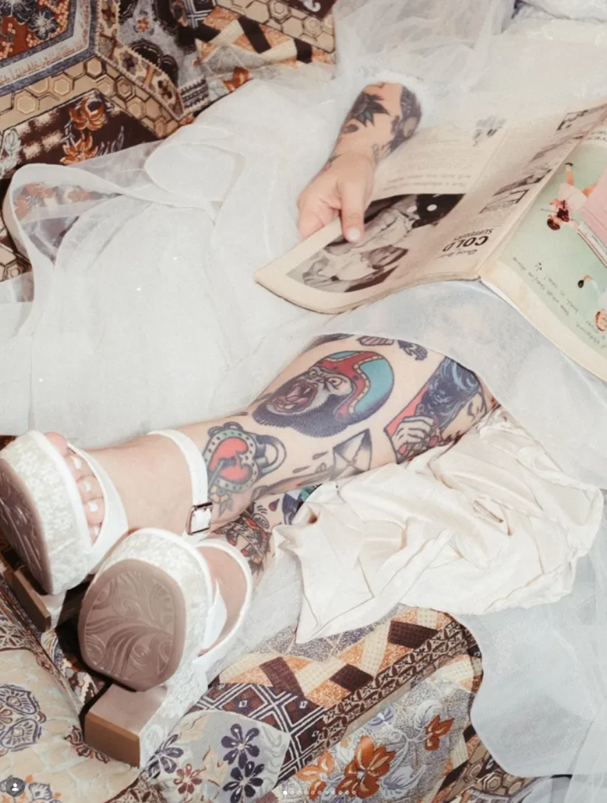A person with tattooed arms and legs, wearing white platform sandals, is lying on a patterned fabric surface, reading a newspaper or magazine.