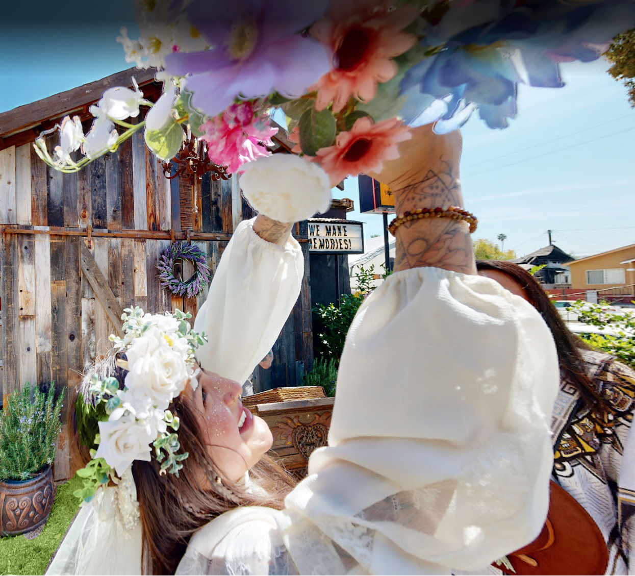 A woman wearing a floral crown catches a bouquet of flowers during an outdoor celebration, with a wooden fence and greenery in the background.